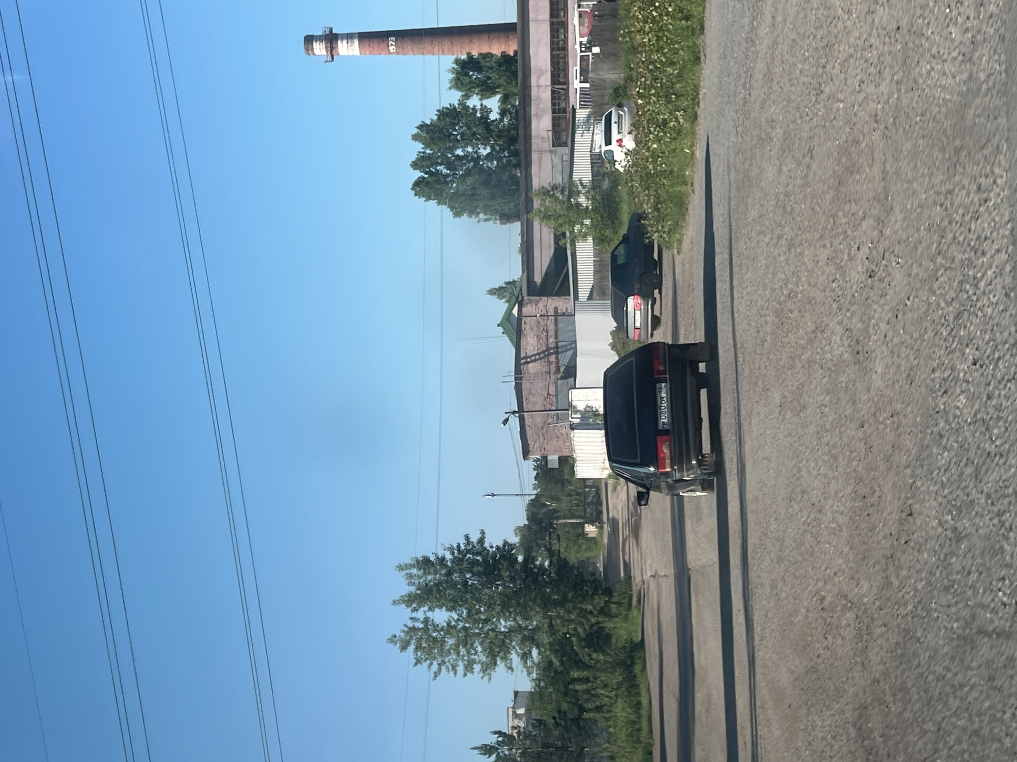 The image depicts a sunlit, outdoor scene in the city of Noviy, Russia. The foreground is a rough, unpaved road. A black sedan is positioned in the center of the road, facing away from the viewer, with its license plate visible. To the right of the black sedan, a white sedan is parked. In the middle ground, there are several buildings, including what appears to be an industrial complex with a large, red brick smokestack that has "1973" written on it. Trees and overgrown vegetation line the sides of the road and surround the buildings. Overhead, power lines are strung across the clear blue sky. The overall impression is one of a somewhat industrial or semi-urban environment under bright daylight. There are no people visible in the image.