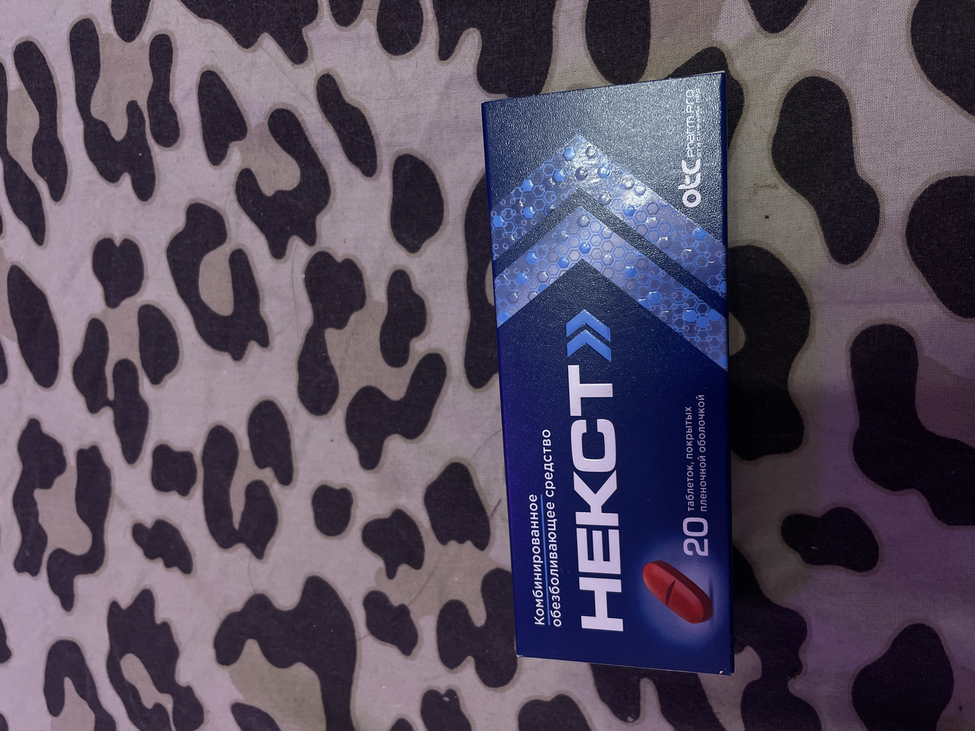 The image shows a close-up, overhead view of a blue box of medication placed on a patterned fabric. The fabric has a camouflage-like pattern with dark and light brown shapes against a lighter background. The medication box is dark blue with white text that reads "Комбинированное обезболивающее средство" (Combined analgesic agent) and "НЕКСТ" (NEXT) in large, bold white letters. Below "НЕКСТ", there is an image of a single red pill, followed by the number "20" and the text "таблеток, покрытых пленочной оболочкой" (tablets, film-coated). In the lower right corner of the box, a logo reads "otc Pharm PRO". There is no visible indication of the location being Vaganovo, Russia, or any people or actions taking place.