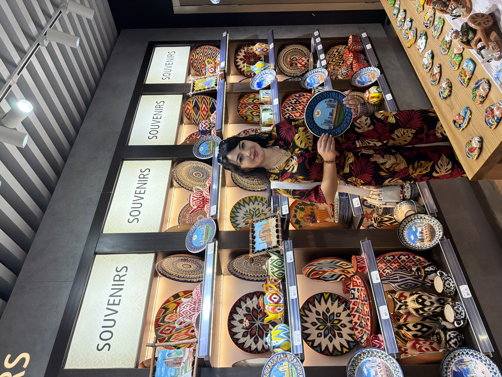 A woman stands in a souvenir shop in Samarkand, Uzbekistan, holding a round ceramic plate. The plate she holds depicts the skyline of Samarkand with the Cyrillic text "Samarkand" below. The shop is filled with shelves displaying various traditional Uzbek handicrafts, including patterned plates, ceramic teapots, vases, and small decorative figures. The woman is wearing a colorful, tropical-print jumpsuit. The environment is well-lit with track lighting.