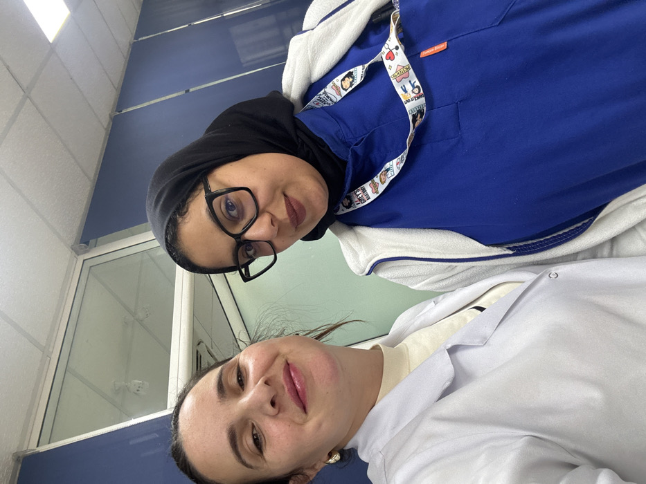 The image displays two adult females in a bright indoor environment, likely a medical or laboratory facility in Meknes, Morocco. The precise location within Meknes is not discernible from the visual evidence. The individual on the left, with dark hair and light skin, wears a white lab coat over a light-colored turtleneck and small gold-colored hoop earrings, looking directly at the camera with a smile. The individual on the right, wearing a black hijab, black-framed glasses, a blue scrub uniform, and a white fleece jacket, also looks forward with a subtle smile. A multi-colored lanyard, featuring graphics and text related to "Grey's Anatomy," hangs around her neck. The background includes white walls, a blue-accented panel or blind, a glass partition, and a suspended ceiling with integrated fluorescent lighting panels.