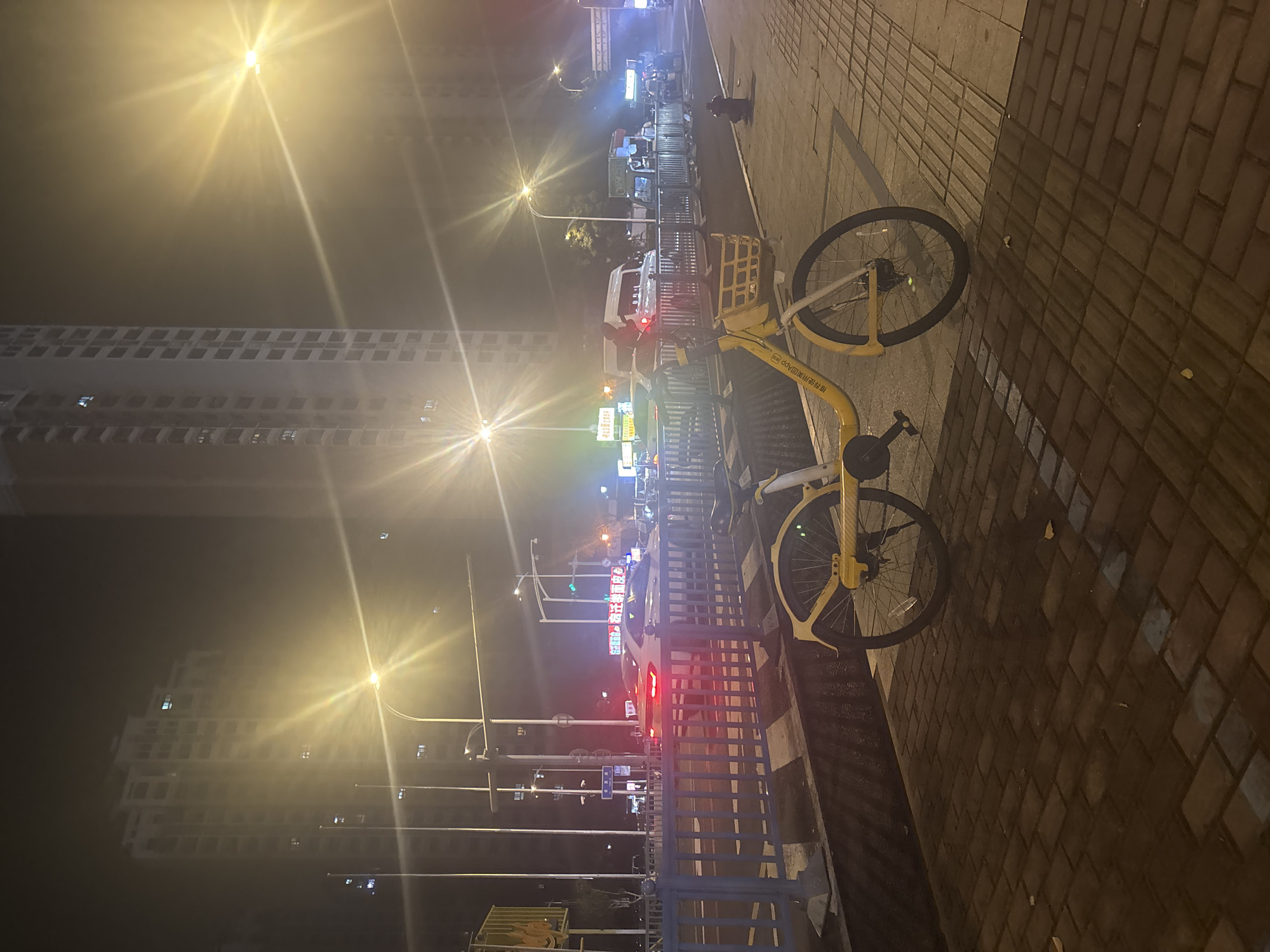 A yellow shared bicycle is parked on a brick sidewalk next to a blue metal fence. In the background, a street with traffic lights and vehicles is visible, with tall apartment buildings and streetlights under a foggy sky. The scene appears to be a nighttime street view in Suzhou Industrial Park, China.