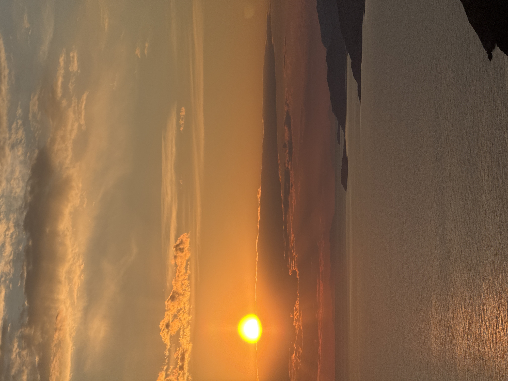 The image shows a sunset in Kovačko Polje, Montenegro. The sun is positioned low in the sky, casting a golden glow across the landscape. To the left of the sun, clouds are visible, illuminated by the sunlight. To the right, land formations and water are visible. The water reflects the sunlight.