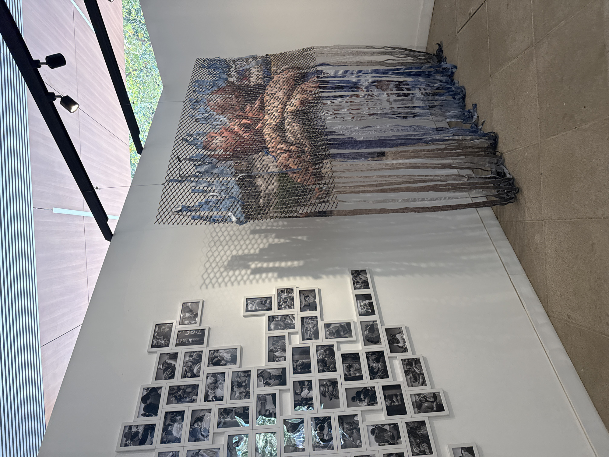 This media file captures an indoor art installation, likely within a gallery or public building in Mexico City, Mexico, during the daytime. The main subject is a white wall displaying two distinct art pieces, with a large window structure to the left and a tiled floor on the right.
The upper artwork is a large, rectangular textile or mixed-media piece, composed of woven or braided strips of fabric in shades of blue, grey, brown, and possibly muted orange or flesh tones. It appears to depict abstract forms or perhaps a human figure, partially obscured or overlaid by a dark, diamond-patterned mesh. Loose fabric strips extend from the bottom right of the piece. A distinct diamond-patterned shadow, cast by a light source from above, is visible on the white wall below this artwork.
Below the textile piece, a dense cluster of framed black-and-white photographs covers a significant portion of the wall. The frames are white and arranged in an irregular, organic pattern. The photographs predominantly feature people, often in close interaction or various poses, though specific details are difficult to discern without closer inspection. One photo frame in the lower section has the text "30-L" visible, and another appears to show "7.0".
To the left of the wall, a large window or glass facade with prominent black vertical supports allows bright natural light to stream in. Lush green foliage is visible outside the window. Black track lighting fixtures with spotlights are mounted on these vertical supports, illuminating the space. The floor is made of large, light-colored square tiles. The scene conveys a contemplative atmosphere, characteristic of an art viewing environment.