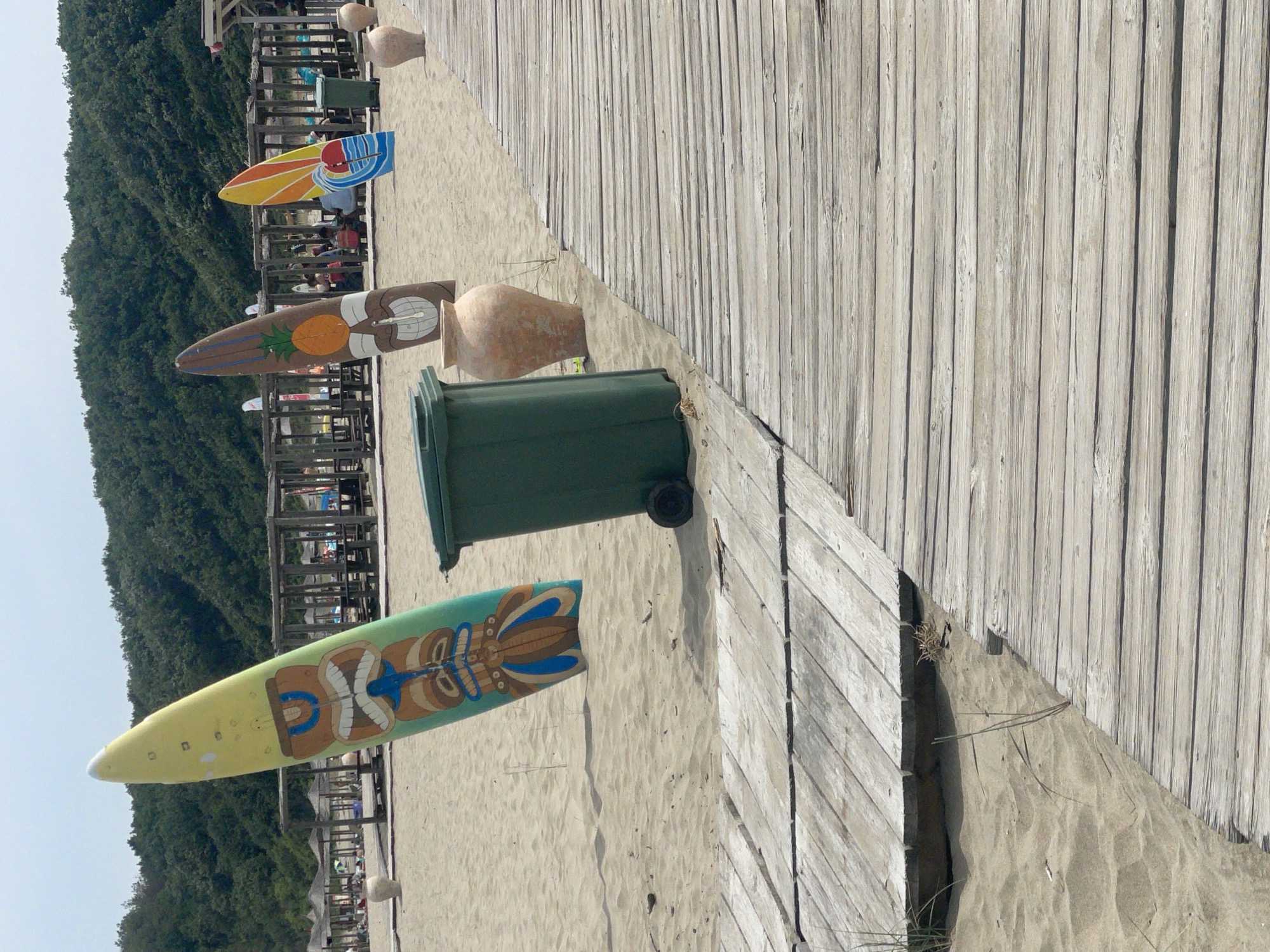 This image depicts a sandy beach in Rezovo, Bulgaria, featuring a wooden boardwalk in the foreground. Three surfboards, decorated with Polynesian-inspired designs, are propped upright in the sand. The first surfboard, on the left, is yellow with a green and brown tiki face. To its right, a brown surfboard displays a pineapple motif. The third surfboard, on the far right, is orange and yellow with a wave design. A green trash bin stands near the middle surfboard, and several large, light-colored amphorae are scattered across the sand. In the background, a line of wooden beach shelters with thatched roofs, umbrellas, and tables and chairs suggests a recreational area. A dense, green forest covers the hillside behind the beach shelters, and the sky above is clear and overcast. The scene is peaceful and appears to be an unpopulated section of the beach.