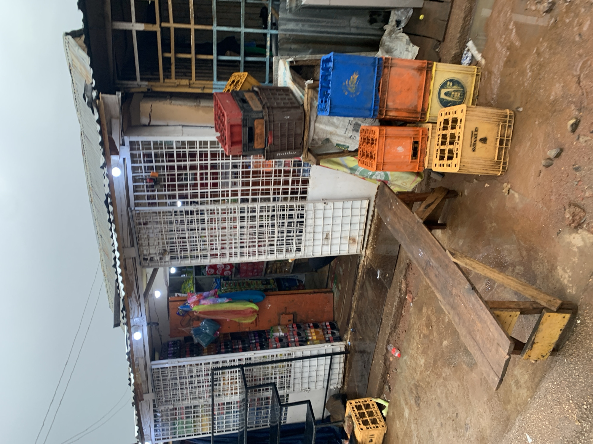 The image shows a small retail shop in New Stadium, Nigeria. The shop is constructed with a corrugated metal roof and a metal frame, caged in with white metal mesh. The interior is stocked with various goods. A collection of plastic crates, stacked in varying colors (blue, orange, brown, and yellow), sits on top of the store's facade. A makeshift wooden structure serves as a bench or counter. The ground outside the shop appears wet, suggesting recent rain.