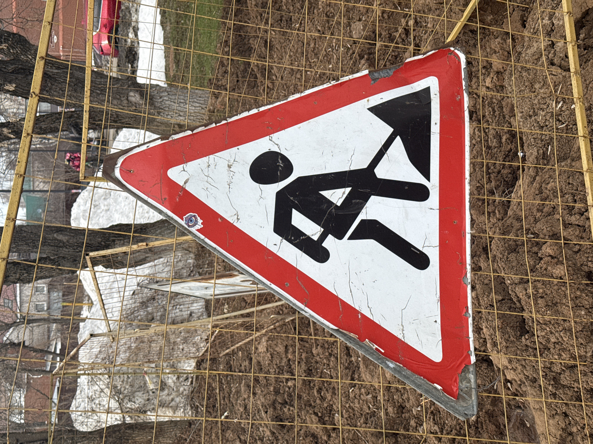 The image shows a triangular red and white road sign with a black silhouette of a construction worker holding a shovel. The sign appears aged and worn, with scratches and faded colors. It is leaning against a yellow wire fence, with dirt and some remnants of snow visible behind the fence. In the background, blurred details of buildings and trees suggest an urban environment. A small sticker can be seen on the sign. The location is Izhevsk, Russia.
