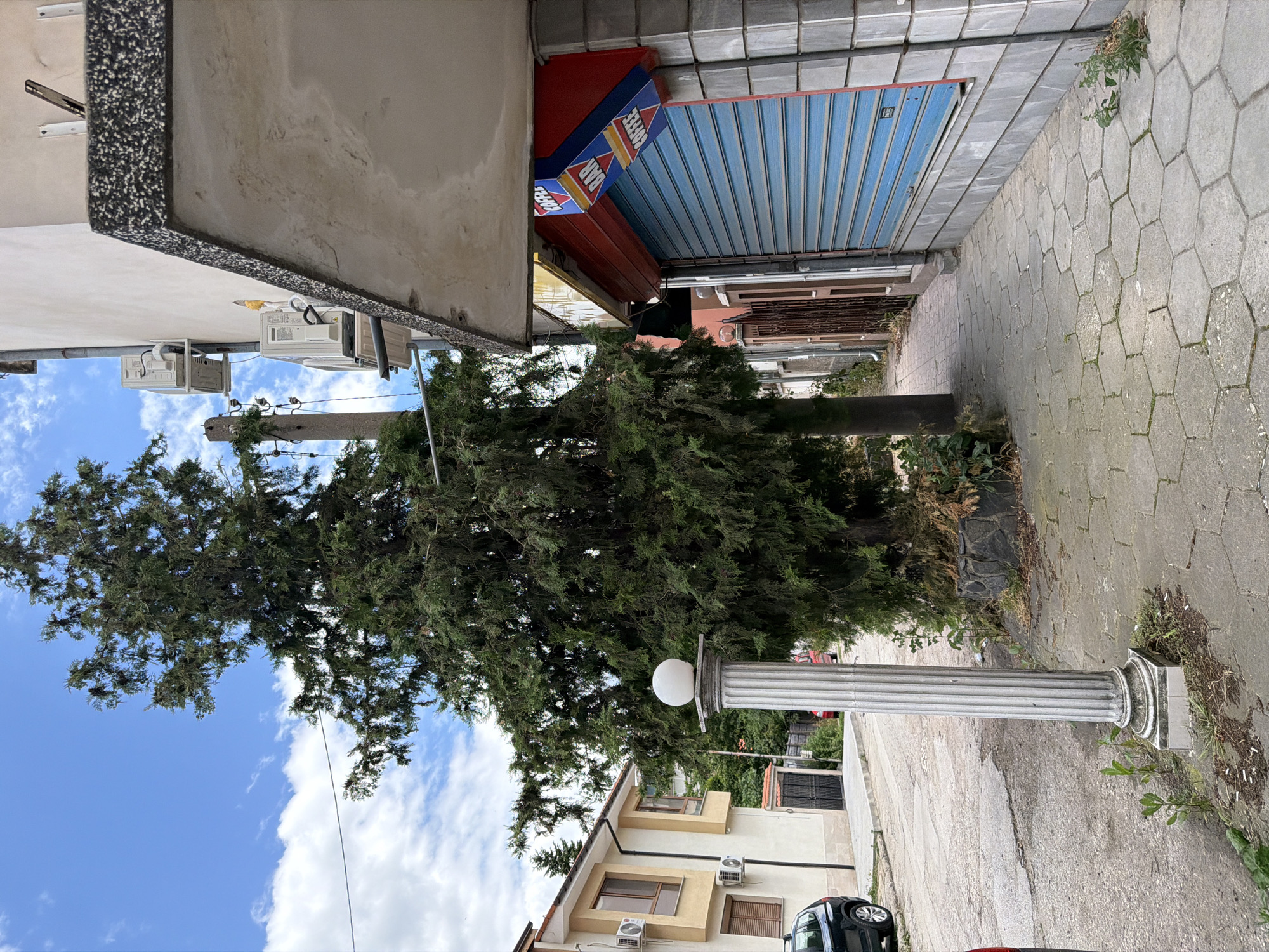 The image shows a street scene in Pazardzhik, Bulgaria, with a focus on a building facade on the right and a large evergreen tree on the left. The building has a blue roller shutter on the ground floor, above which is a sign for "COFFEE BAR". Several air conditioning units are mounted on the exterior wall of the building to the left of the shop. A white, fluted column with a spherical light fixture sits on a paved sidewalk in front of the tree. The street is paved with asphalt and the sidewalk is made of hexagonal concrete tiles. A black car is parked on the left side of the street. A residential building with cream-colored walls and brown window frames is visible in the background on the left. The sky is blue with scattered white clouds.