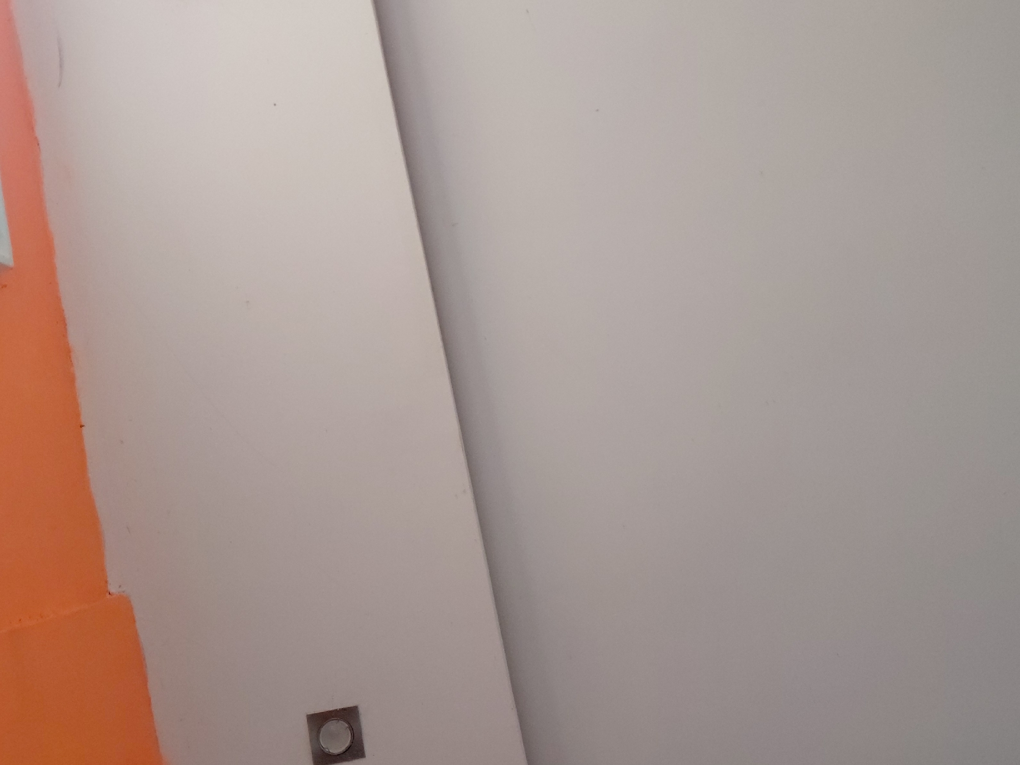 The image displays an indoor wall featuring a distinct color scheme and a small architectural detail. The left portion of the wall is painted a vibrant orange, while the larger right section is a light, neutral off-white or very pale grey. A sharp, diagonal line divides these two painted areas, suggesting a corner or an intentional design element within a room.
In the lower-left area of the neutral-colored wall, there is a small, square-shaped metallic object. This object has a circular element, possibly a button, indicator light, or sensor, positioned centrally within its square frame. No people or overt activities are visible in the frame. The lighting appears even, suggesting either diffused natural light or interior artificial lighting, making the time of day indeterminate. There are no visible textual elements. While the location is specified as Santa Ana, Venezuela, no specific visual cues within the image confirm this setting.
