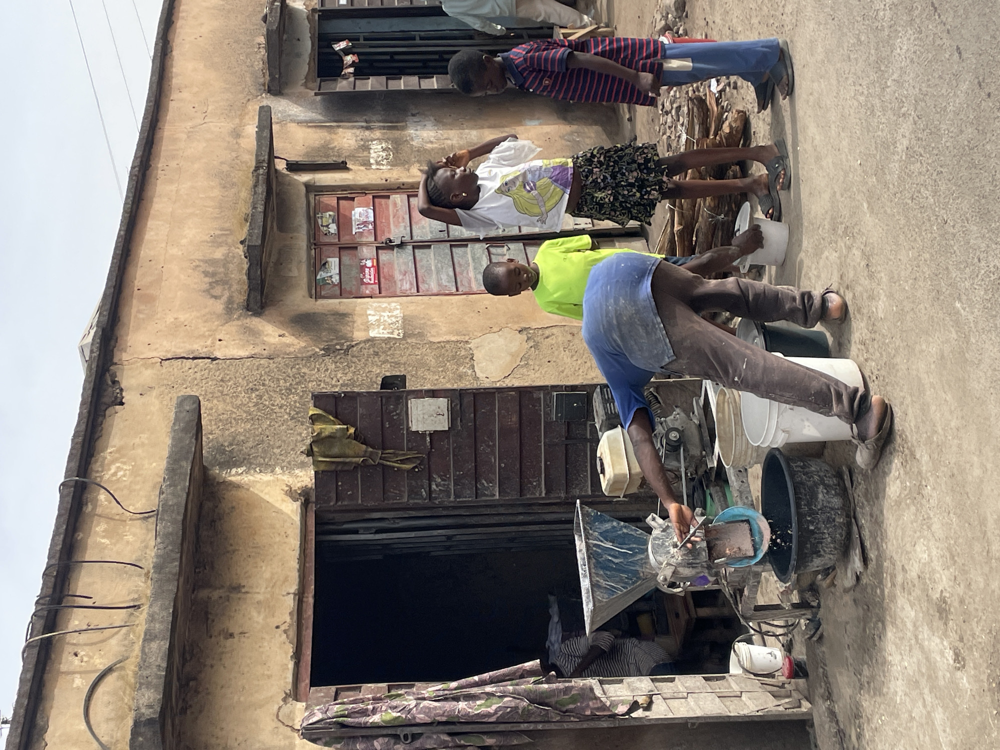 This image, rotated 90 degrees clockwise, depicts an active street scene in Jos, Nigeria, in front of an aged, multi-story building under an overcast sky.
In the foreground, a dark-skinned man wearing a blue t-shirt and brown pants is operating a mechanical grinding machine. He is bending over the equipment, which has a metal hopper and is surrounded by several white and black plastic buckets and tubs. Material is visibly being processed and collected into one of the containers.
Standing behind the man, to the left (when viewed correctly), are three children. A young boy in a bright neon green t-shirt is partially visible, looking towards the machine. Beside him, a girl with dark skin wears a white t-shirt featuring a Disney princess (Rapunzel) and a patterned skirt. She has an earring and holds her hands near her head as she observes. Further back, another boy, also with dark skin, is seen in a red and blue striped t-shirt and blue pants, looking down.
The background is dominated by a dilapidated building with light brown, weathered walls showing cracks. The lower section features a dark, open entryway. Above it, a set of closed, reddish-brown double doors is adorned with small, faded posters or flyers; one suggests a "Coca-Cola" branding, but is largely illegible. Higher up, a window is partly covered by a draped yellow/brown fabric, and exposed rebar is visible on the building's exterior. The ground is a worn, concrete-like surface. The bright, diffused light indicates it is daytime. The scene portrays a slice of daily life, possibly a small street-side business, with children nearby.