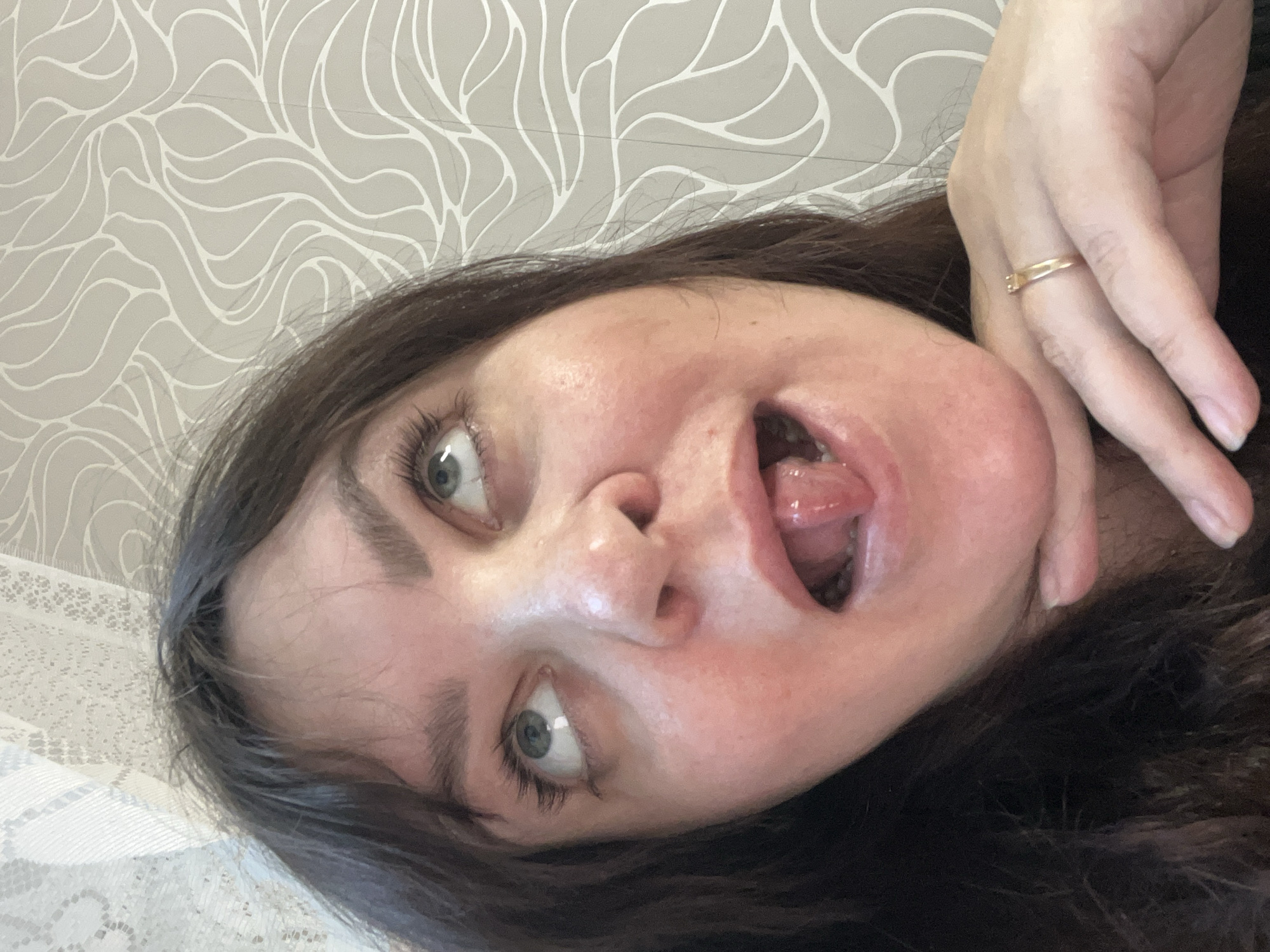 A close-up, sideways portrait captures a woman with dark hair and striking green-blue eyes. Her face is presented in an exaggerated expression, with her mouth wide open and tongue partially extended, while her eyes gaze upwards and to the left. The overall demeanor suggests playfulness or a moment of surprise. Her fair-skinned right hand, adorned with a simple gold band ring, is visible on the right side of the frame, resting near her dark hair. The background consists of light grey wallpaper patterned with white, abstract, wavy leaf-like motifs. In the lower-left corner, a patch of white lace fabric, possibly from a curtain or tablecloth, is visible. The setting is an informal indoor space, implied to be a room in Perm, Russia. No text is visible within the image.