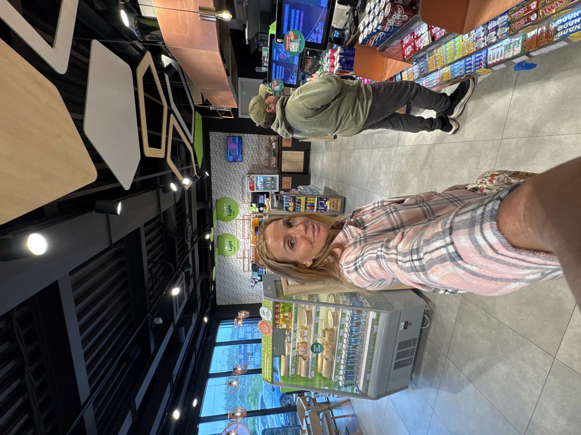 The image shows two people inside a brightly lit convenience store or a small supermarket. A woman in a pink and gray plaid shirt is in the foreground, looking towards the camera with a slight smile. Behind her, a man wearing a green hooded jacket and a green baseball cap is standing at a checkout counter, holding a phone. The store shelves are stocked with various products, including drinks, snacks like Twix and Bounty bars, and some refrigerated items. The overall atmosphere appears casual and everyday.
Visible text includes "Green Light" and "Red Bull" branding, as well as some Russian text at the checkout counter which seems to relate to fuel types and prices, suggesting the store might also function as a gas station convenience store. The lighting is artificial and bright, indicating it's likely daytime. The flooring is tiled, and the ceiling features industrial-style lighting and exposed ductwork. The setting appears to be a modern retail environment.