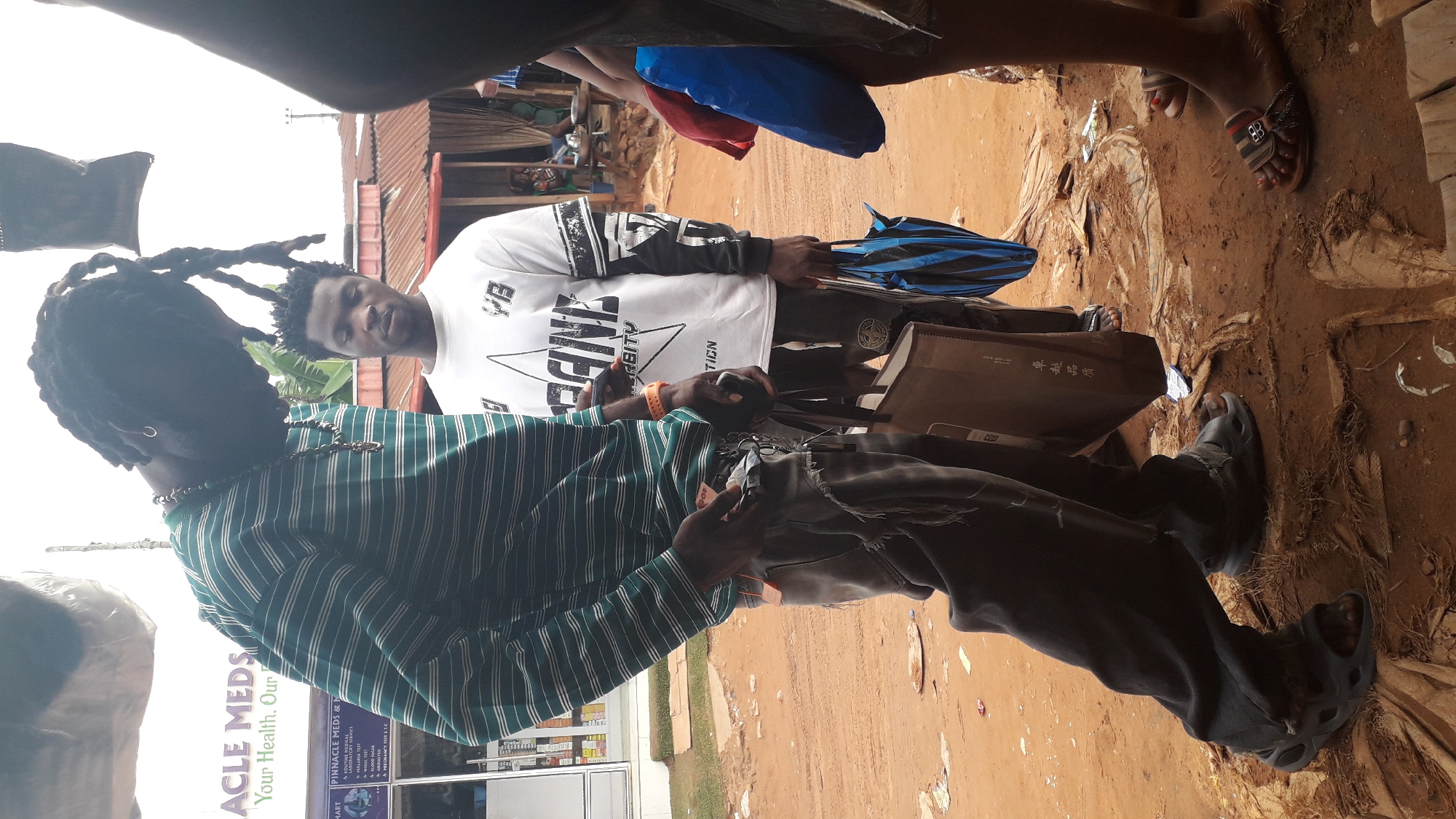 The image shows two men in Nkpor, Nigeria, standing outdoors. The man in the foreground wears a green and white striped shirt, a necklace, and black pants with Crocs-like shoes. He has dreadlocks. The other man is wearing a white shirt with black sleeves and lettering. He is holding a folded blue umbrella and a shopping bag. The ground is dirt and trash. Behind the men is a building with signage visible that reads "ACLE MEDS Your Health, Our" and "PINNACLE MEDS &". There are various product signs visible in the building's windows.