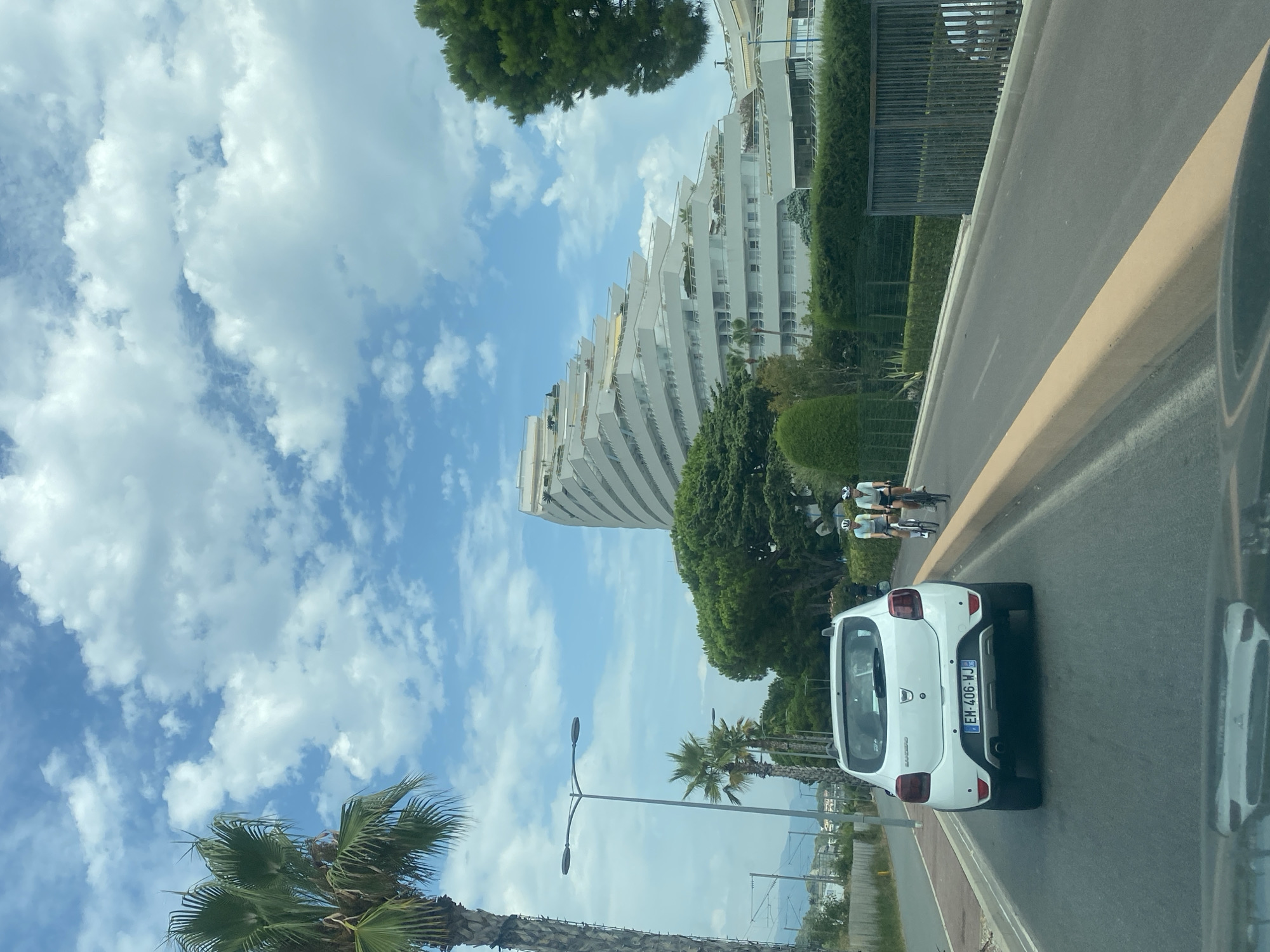A white Dacia Sandero is driving on a road in Villeneuve-Loubet, France. Two cyclists, a man and a woman, are riding in the same direction in front of the car. In the background, there is a distinctive tiered white building with balconies, characteristic of the architecture in Villeneuve-Loubet. Palm trees and other lush green vegetation line the road and surround the building. The sky is blue with fluffy white clouds, indicating a sunny day.