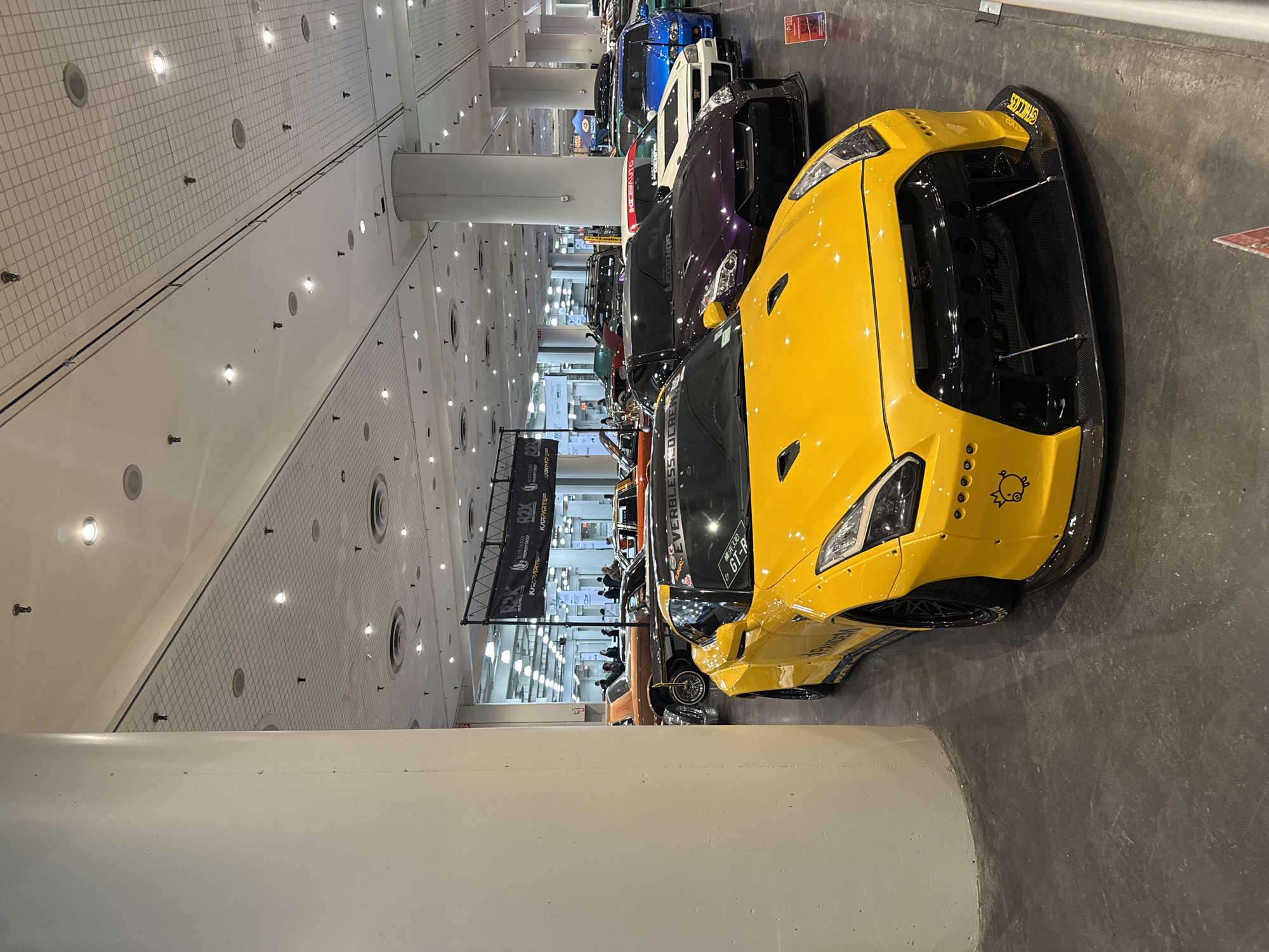 The image shows a yellow Nissan GT-R sports car on display at what appears to be an indoor car show or exhibition. The car is prominently featured in the foreground, parked on a grey concrete floor. Other cars are visible in the background, including models in purple, white, and blue. Several banners with logos and text are visible behind the cars, indicating sponsors or exhibitors. The lighting is bright and even, suggesting a well-lit indoor space. The environment seems to be a large convention center or exhibition hall with a high ceiling and numerous lights. Given the presence of the vehicles and the overall layout, it is likely in New York City.