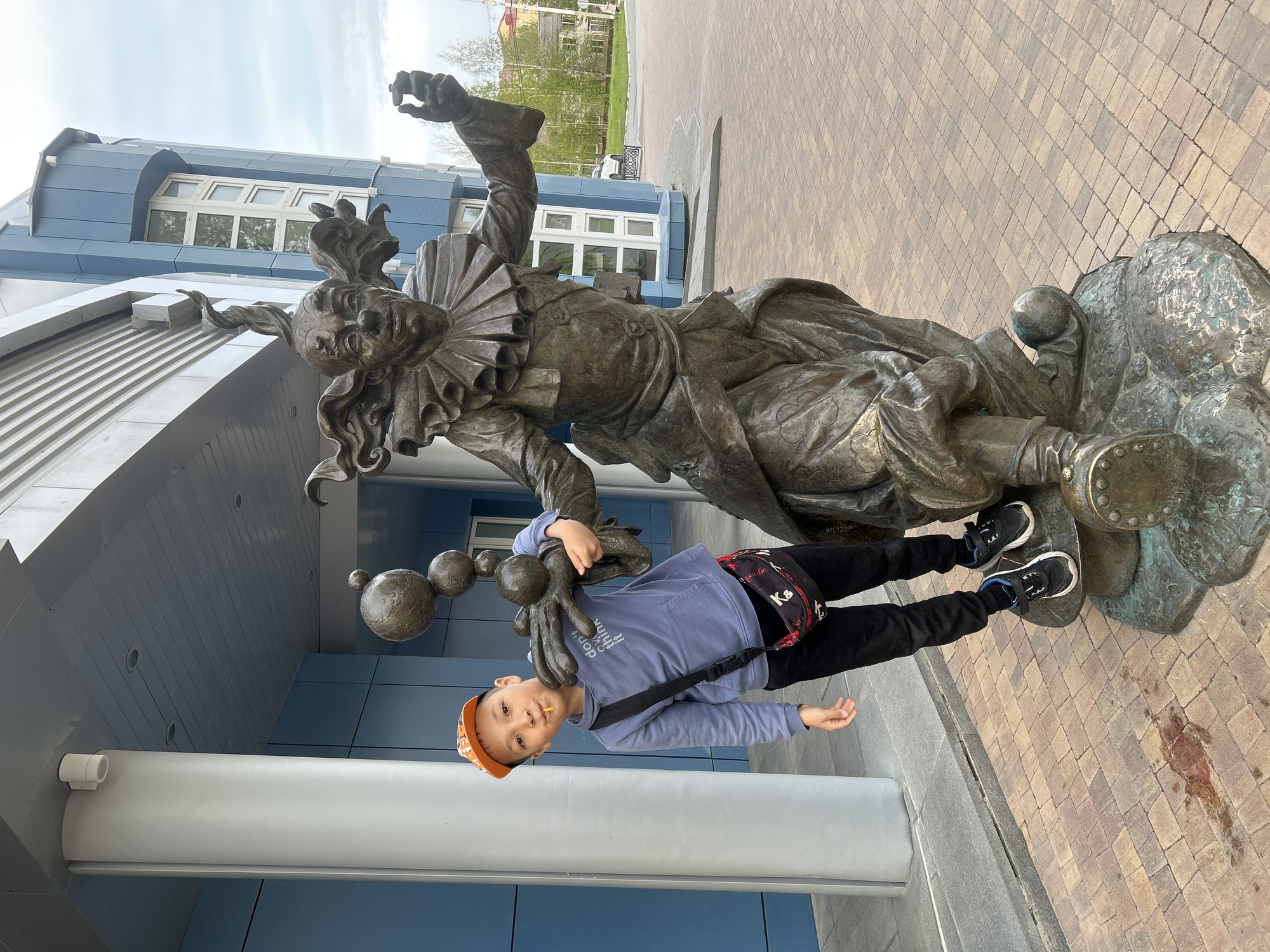 A young boy, wearing an orange cap, light blue sweatshirt, black pants, and black sneakers, is posing next to a bronze sculpture in what appears to be Uray, Russia. He has a black messenger bag across his body. The sculpture depicts a figure in a theatrical costume with a ruffled collar, seated on a cloud-like base. Ornate spheres are connected to the left side of the statue. The boy has his left hand holding onto one of these spheres. They are positioned on a paved walkway made of light brown brick tiles. Behind them is a modern, light blue building with visible windows and a white supporting column. The sky is overcast and bright. The building behind them has a sign that reads "41012-06" in white text on a black sign. Another building with red accents, trees, and greenery is visible in the distance. There is a dark stain on the brick tiles next to the base of the statue.