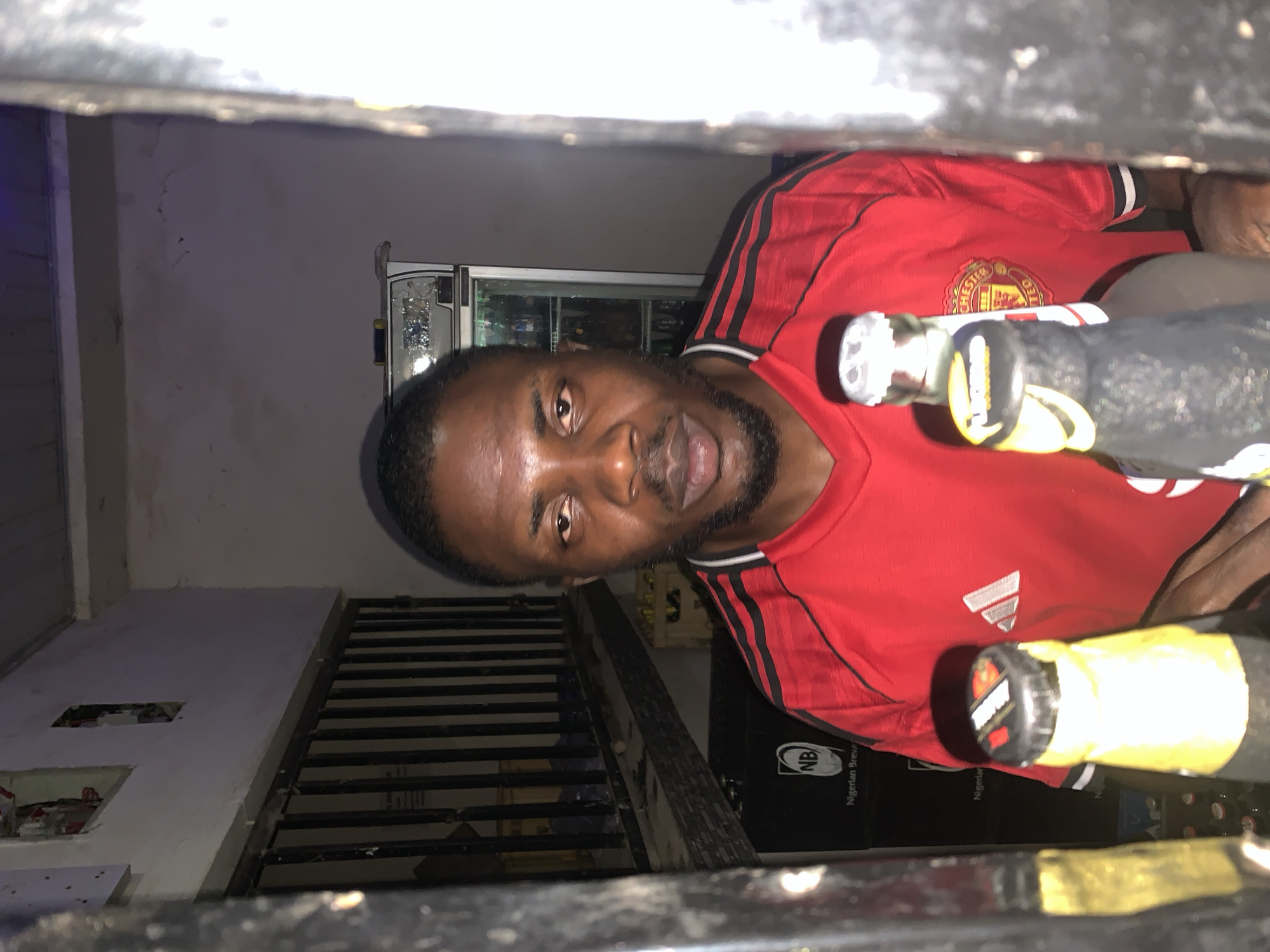 A man wearing a red Manchester United jersey is seen indoors. He appears to be holding bottles, possibly alcoholic beverages. In the background, there is a refrigerator and a black container with the inscription "NB Nigerian Breweries." The location is potentially a bar or retail establishment in Kabong, Nigeria. The viewpoint is from behind a dark, possibly metal, structure.