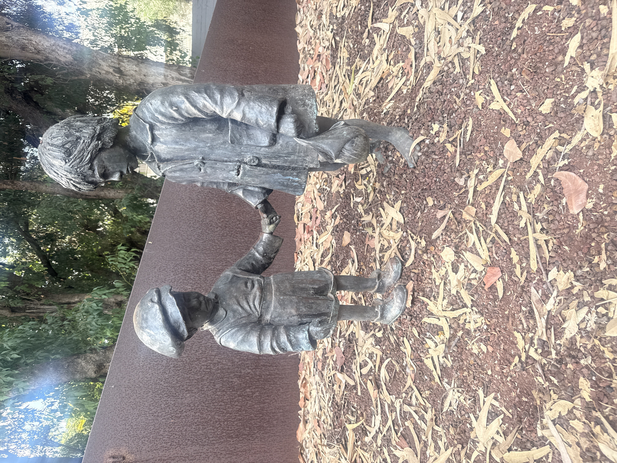 This is a full-shot, eye-level, outdoor photograph of a bronze sculpture depicting two young children holding hands. The sculpture is situated in a public space, possibly a park or memorial, with a rough, rust-colored metal wall behind it and a ground covered in reddish-brown gravel and dry, fallen leaves.
The child on the left is shorter and wears a cap and what appears to be a romper-style outfit with suspenders. This child is looking down. The child on the right is taller and wears a coat with buttons and a collar. This child is holding a bundle or bag in their left hand and is looking down and slightly to the side. Both children appear to be barefoot. The overall impression is one of innocence and perhaps a shared moment of vulnerability or adventure. The lighting suggests it is daytime, with sunlight filtering through the trees in the background, casting dappled light and shadows. The trees in the background are large and leafy, adding to the natural setting.
This sculpture is located in Ramat HaSharon, Israel, specifically in the Rabin Memorial Park. It is part of the "Children of the World" exhibit, a collection of sculptures donated by various countries to commemorate children lost to war and terrorism. This particular sculpture, titled "The Orphans" or "The Wanderers," represents children who have lost their families.