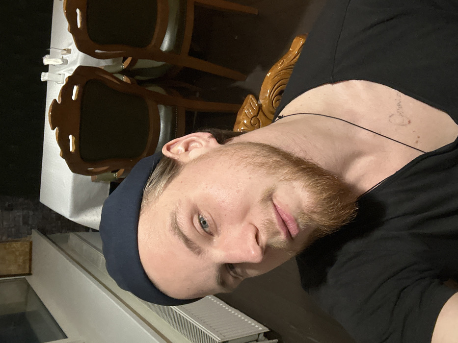 A man with a ginger beard and blue eyes wears a dark blue beanie. He's wearing a black t-shirt, and a thin black cord necklace. An ornate wooden chair is behind him, near a white tablecloth and chairs. Part of a radiator is visible. He appears to be indoors with dim lighting.