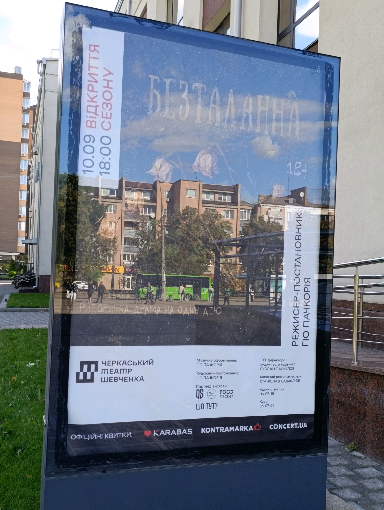 A vertical, outdoor advertisement is displayed on a black stand in front of a building in Cherkasy, Ukraine. The advertisement is for the Cherkasy Academic Music and Drama Theater named after Taras Shevchenko. The poster, protected by glass, features a large white text overlay on a reflective surface showing a reflection of a street scene with buildings, trees, and people. The title of the performance appears to be "BEZDALANNA". Smaller text details include the opening date and time (10.09, 18:00), and the genre (Rhetorical Drama in One Act). Information about the theater, production staff, and ticket vendors (Karabas, Kontramarka, Concert.ua) is also present at the bottom. The scene is set on a sidewalk with green grass to the left and a modern building with metal railings to the right.
