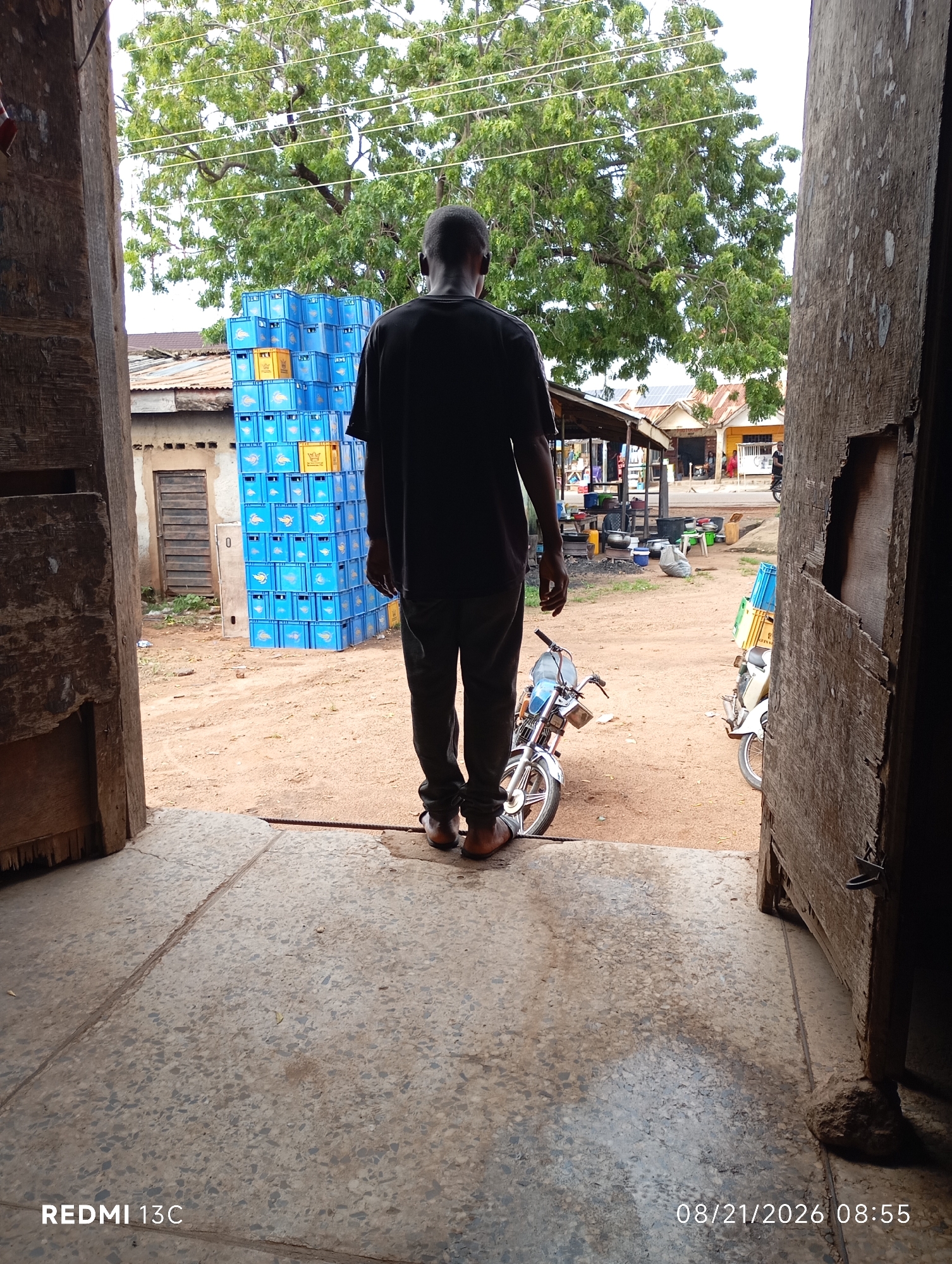 A man stands facing away from the camera, viewed from the doorway of a building, looking out onto a dirt yard. He is wearing a black t-shirt and dark trousers. To his left, stacked high, are numerous blue crates, some yellow crates visible within the stack. In front of the man and slightly to his right is a white and blue motorcycle. In the background, beyond the yard, are several buildings with corrugated metal roofs. The ground is composed of concrete in the foreground and dirt in the yard. The image is dated 08/21/2026 at 08:55. This scene is located in Shendam, Nigeria.