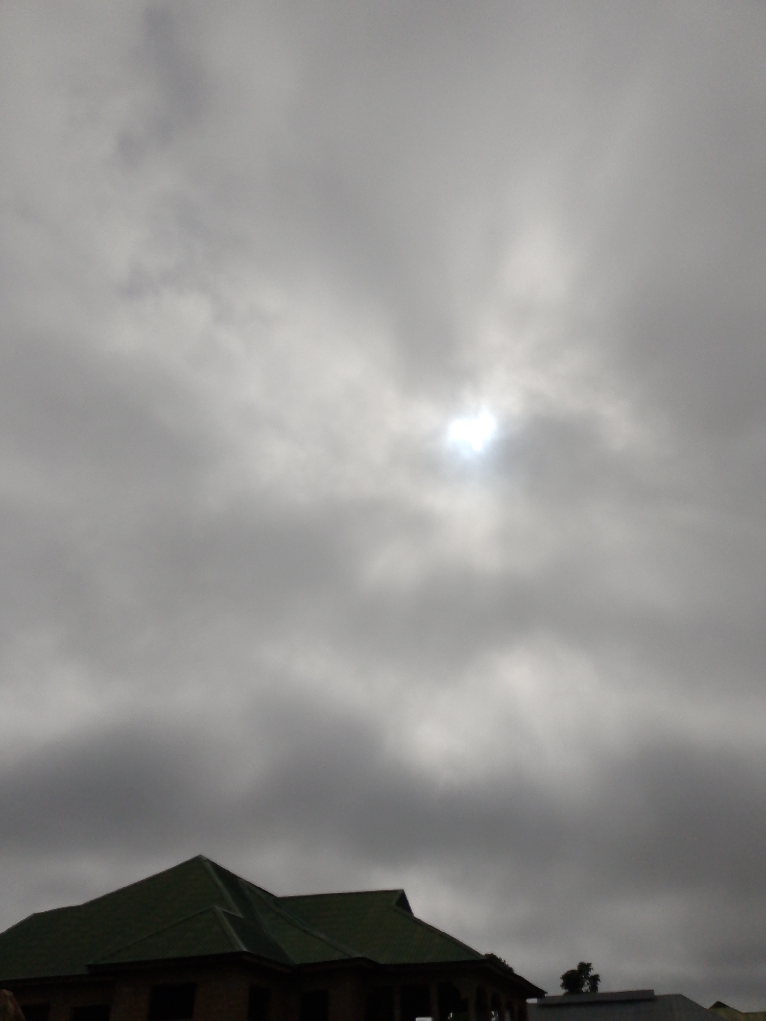 The image shows a building with a green roof under a heavily overcast sky. The sun is visible as a bright spot through a break in the clouds. The location is likely Barikin Gidar Kanat, Nigeria. Other buildings are visible in the background. No people are seen.