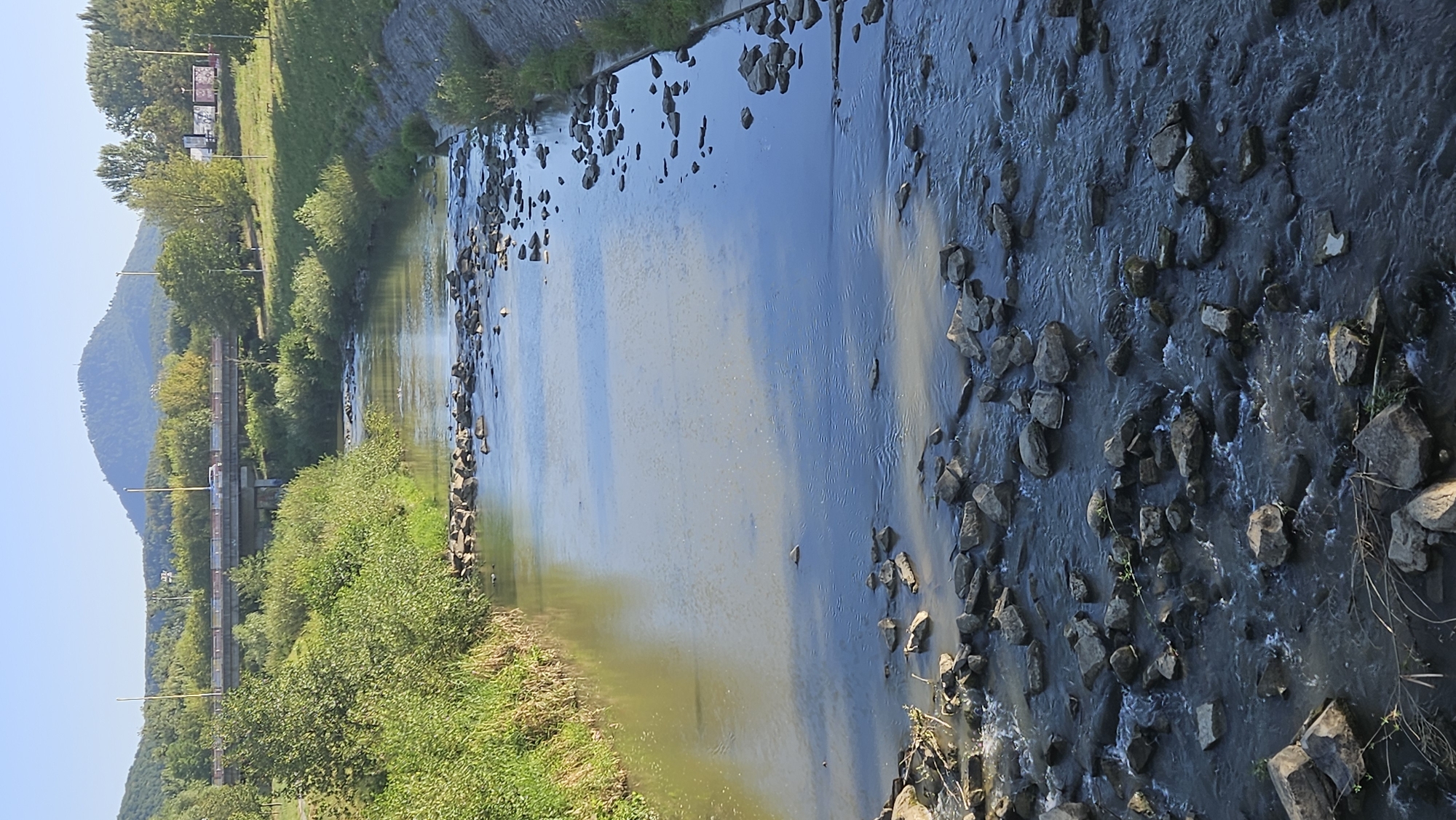 The image depicts the Torysa River flowing through a park-like area in the city of Prešov, Slovakia. The river is partially lined with concrete retaining walls and filled with rocks of various sizes. Lush green vegetation grows along the riverbanks, and in the background, a large hill or mountain with a small structure at its summit dominates the skyline. A bridge with vehicles crossing it is visible in the middle distance, suggesting urban infrastructure nearby. The sky is clear and blue, indicating a sunny day.