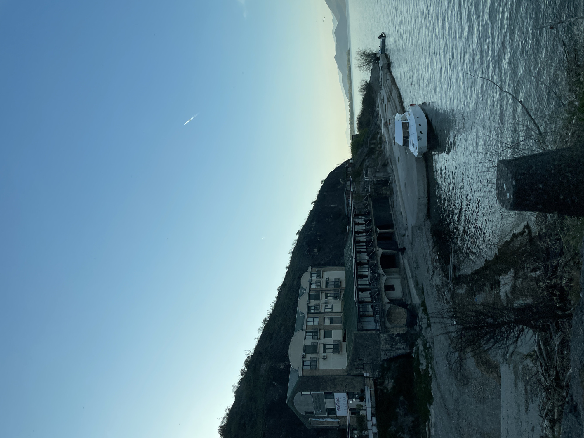 The image shows a waterfront scene in Kruševica, Montenegro. In the foreground, there is a gray concrete wall, followed by a road. A white boat is docked along the concrete pier. A solitary figure stands at the end of the pier near the water. Behind the road, a building is built into the slope of a hill. The building appears to be a multi-story hotel with several visible windows and a sign that says "Jezero." The hill is covered with dark green vegetation. The sky is a clear gradient of light blue. A contrail stretches across the sky. In the background, more hills are visible across the water. The water is calm.