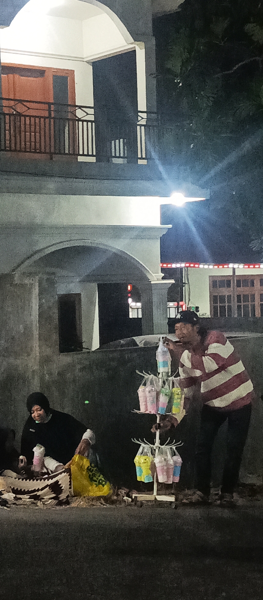 A group of people are gathered on a sidewalk at night in front of a building. The building appears to be a residential structure with an arched entranceway and a balcony with railings on the upper floor. A person in a black headscarf and black top sits on the ground, holding a pink cup and a yellow bag. To their right, a person wearing a striped shirt and a baseball cap stands next to a display rack holding several colorful drinks in plastic bags. The ground is paved and appears wet. Streetlights illuminate the scene, casting shadows and creating a somewhat grainy image. The overall atmosphere suggests a street-side vendor operation in a city or town.