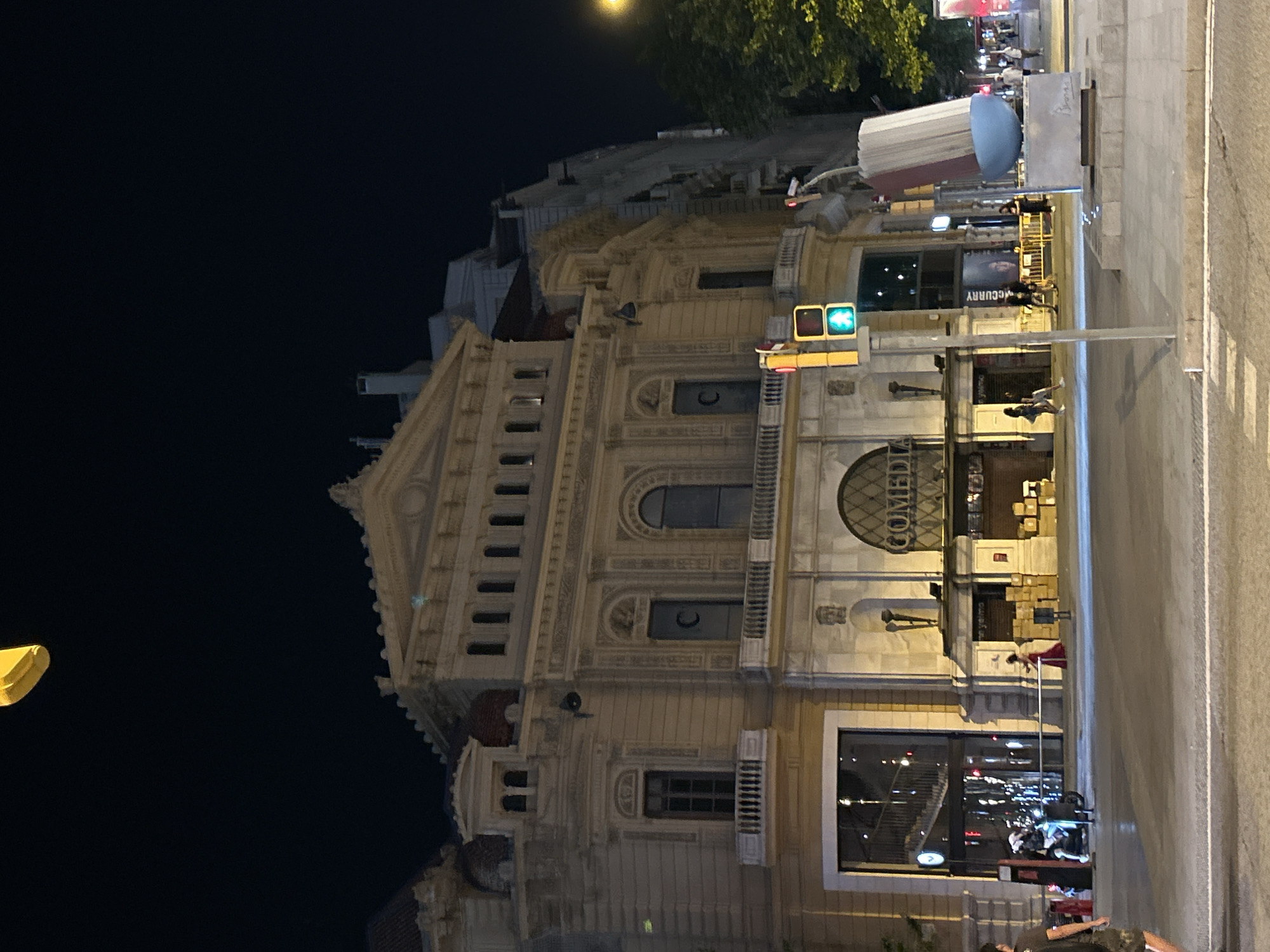 The image is of the Teatre Comedia in Barcelona, Spain, taken at night. The building is illuminated and the green light of a traffic signal is seen to the left of the facade. There are people standing on the sidewalk in front of the building. A large advertisement banner with “McCurry” on it is visible. A street pole is in front of the building and the edge of a street is on the right.