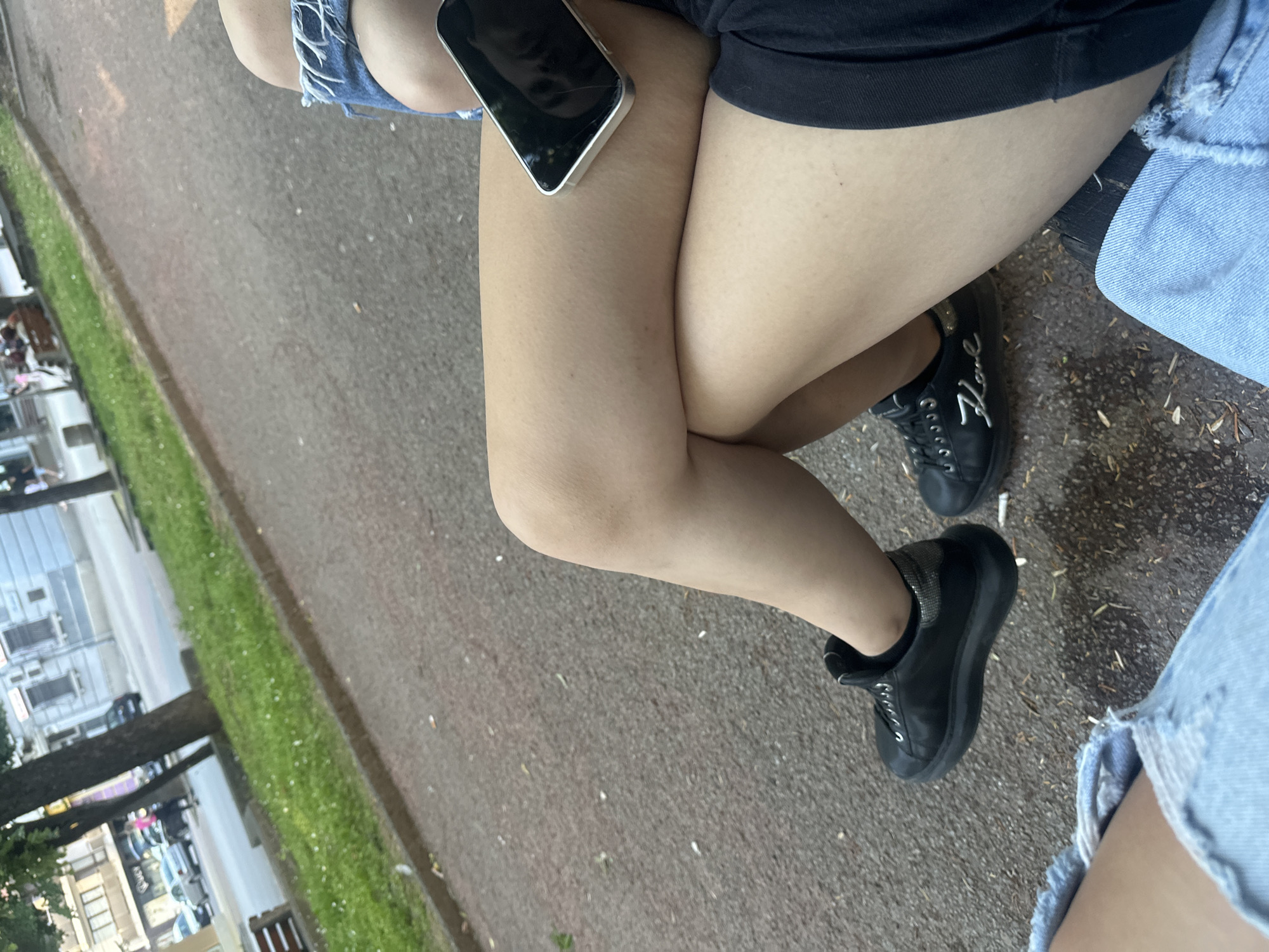 The image depicts a close-up view of two individuals sitting outdoors, likely on a bench, with their legs prominently displayed. The ground appears to be a paved surface, possibly a walkway or park path, with scattered debris. In the background, a street scene is visible with buildings, trees, and parked cars, suggesting an urban setting. The individuals are wearing casual clothing, including denim shorts and black sneakers. One individual's hand is holding a smartphone. The lighting suggests it is daytime. The overall scene conveys a relaxed, everyday moment. Based on visual cues, such as the architecture and vegetation, the location is consistent with a park or public space in Shumen, Bulgaria.