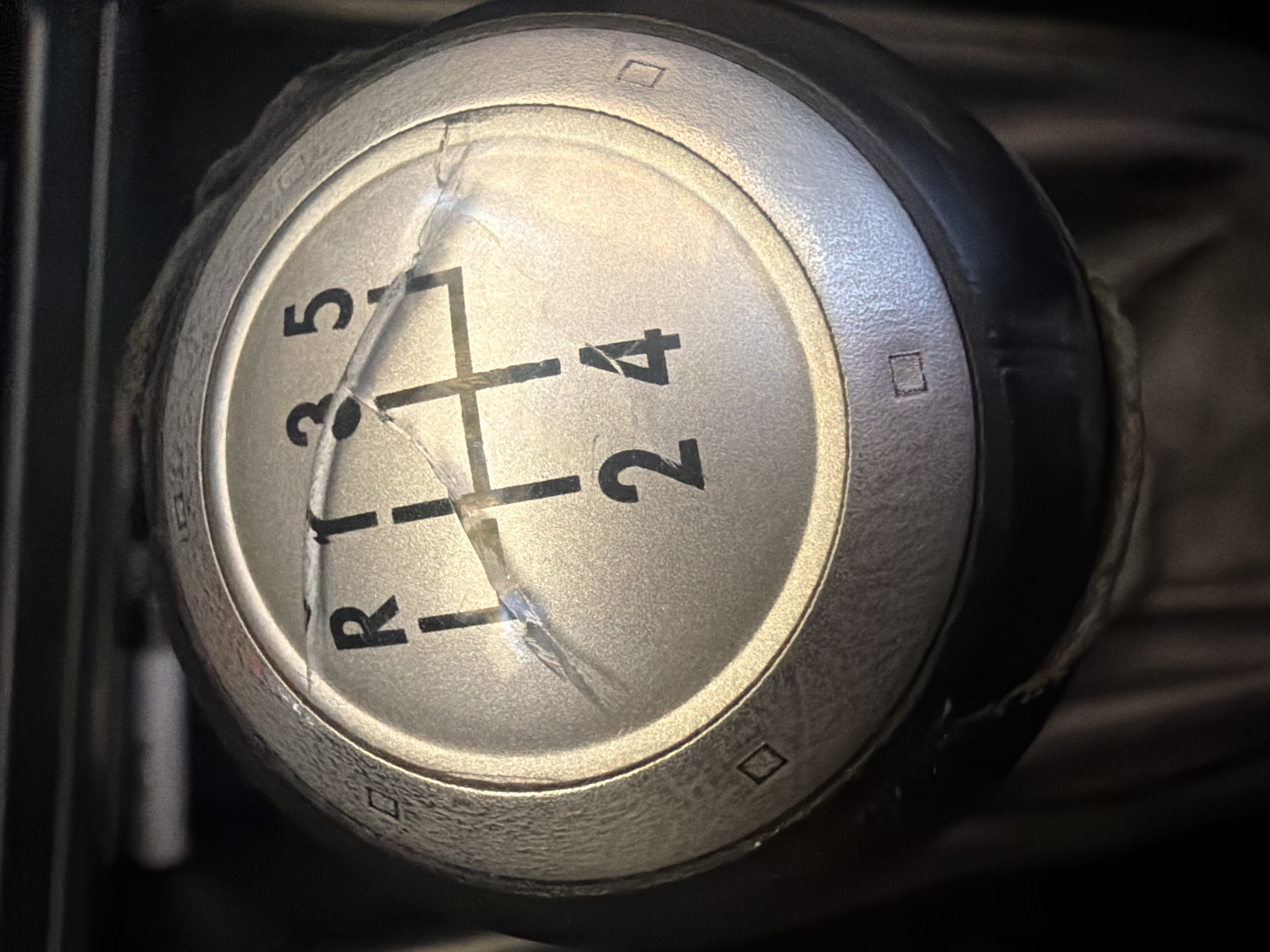 The image shows a close-up view of a gear shift knob, likely from a manual transmission vehicle. The knob is silver with black lettering indicating the gear positions: R (reverse), 1, 2, 3, 4, and 5. A schematic shows where each gear is located. There are cracks on the top surface of the knob. The knob has a black leather boot around its base. The photo is taken in a dark environment. There is no indication of this image being taken in Dolna Mitropolia, Bulgaria. There are no people, actions, or interactions shown.