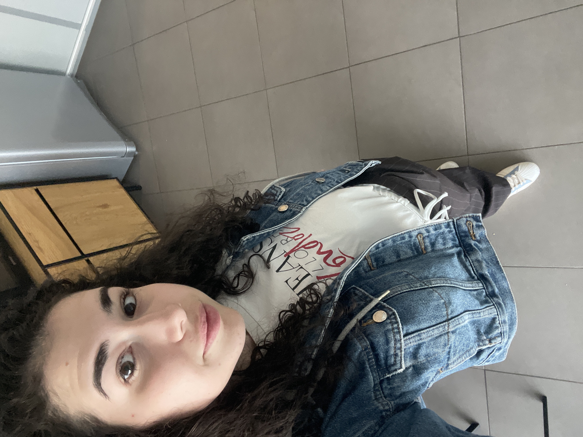 A young woman with long, dark, curly hair lies on a tiled floor indoors, looking up at the camera with a neutral to slightly smiling expression. She is wearing a white t-shirt under a blue denim jacket, paired with dark pinstriped trousers. One light-colored sneaker, possibly white with light blue accents, is visible on her right foot.
The setting appears to be indoors, with the floor covered in large, light gray square tiles. In the background, to the woman's left, a metallic gray appliance or piece of furniture is visible, next to a wooden surface. The lighting is soft and even, typical of an indoor environment, with no direct sunlight or explicit time of day cues.
On her white t-shirt, visible text in both red and black is present, partially obscured by her hair and jacket. The red text appears to include "Lover" or part of it, while black text resembling "JEANS" is also discernable. The image is rotated 90 degrees counter-clockwise. The location context provided is Sofia, Bulgaria, though this is not visually evident in the image itself.