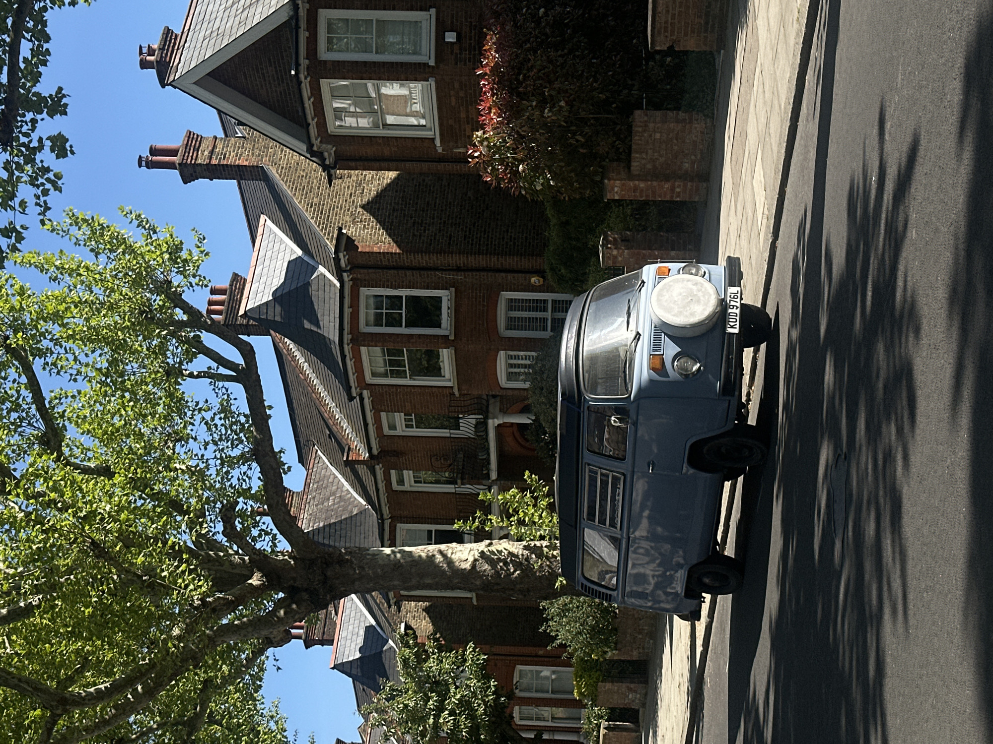 A vertical, medium shot depicts a blue Volkswagen camper van parked on the street in a residential area. The van has a spare tire mounted on the front. Behind the van are several brick houses with tiled roofs and chimneys. A large tree with green leaves is on the left side of the frame. The asphalt road is visible in the foreground, with tree shadows. The license plate is KOD 976L. The location may be in London, UK, given the architecture and license plate style.