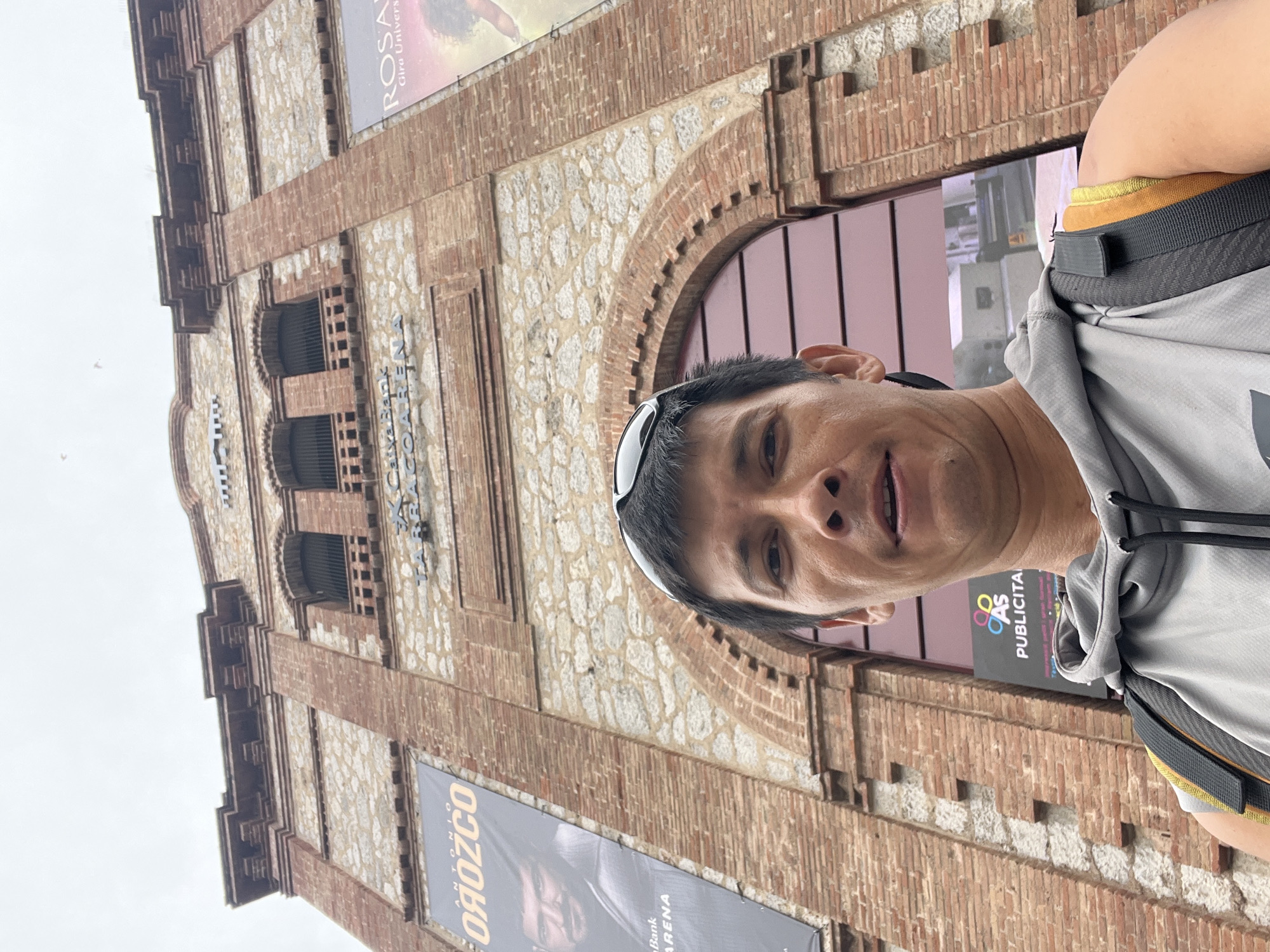 The image is a low-angle, eye-level selfie taken by a man in front of the Palau d'Esports Catalunya in Tarragona, Spain. The building, constructed of red brick and stone, features a large arched entrance and prominent banners advertising "ANTONIO OROZCO" and "ROSANA". The man, wearing a grey hoodie and a backpack, is looking directly at the camera. The sky is overcast.