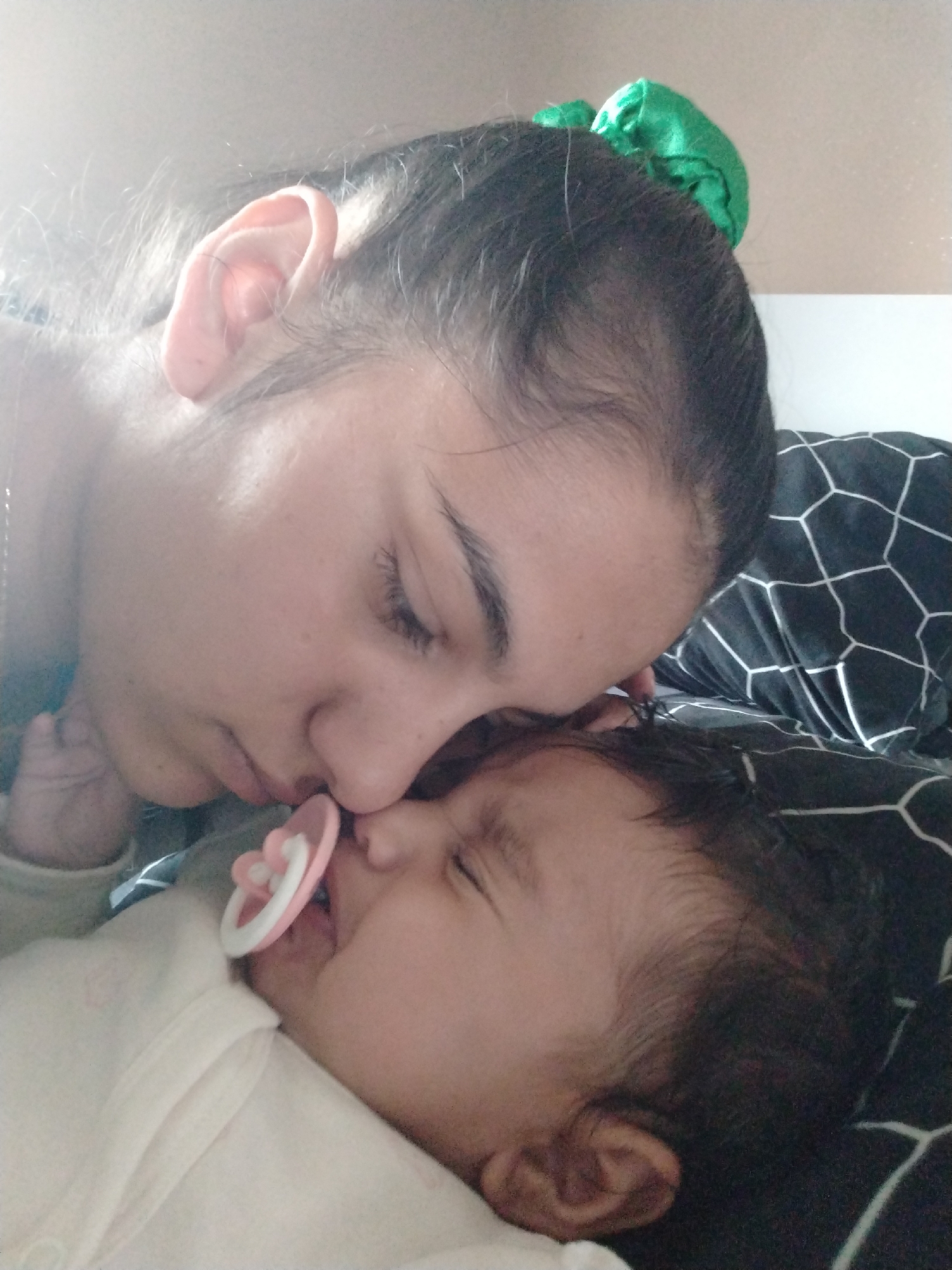 The photograph depicts a woman and a baby lying close together on a bed. The woman has dark hair pulled back with a green scrunchie and is resting her face near the baby's. The baby is lying on its back, eyes closed, with a pacifier in its mouth. The bedding is a black and white geometric pattern. The location is unknown.