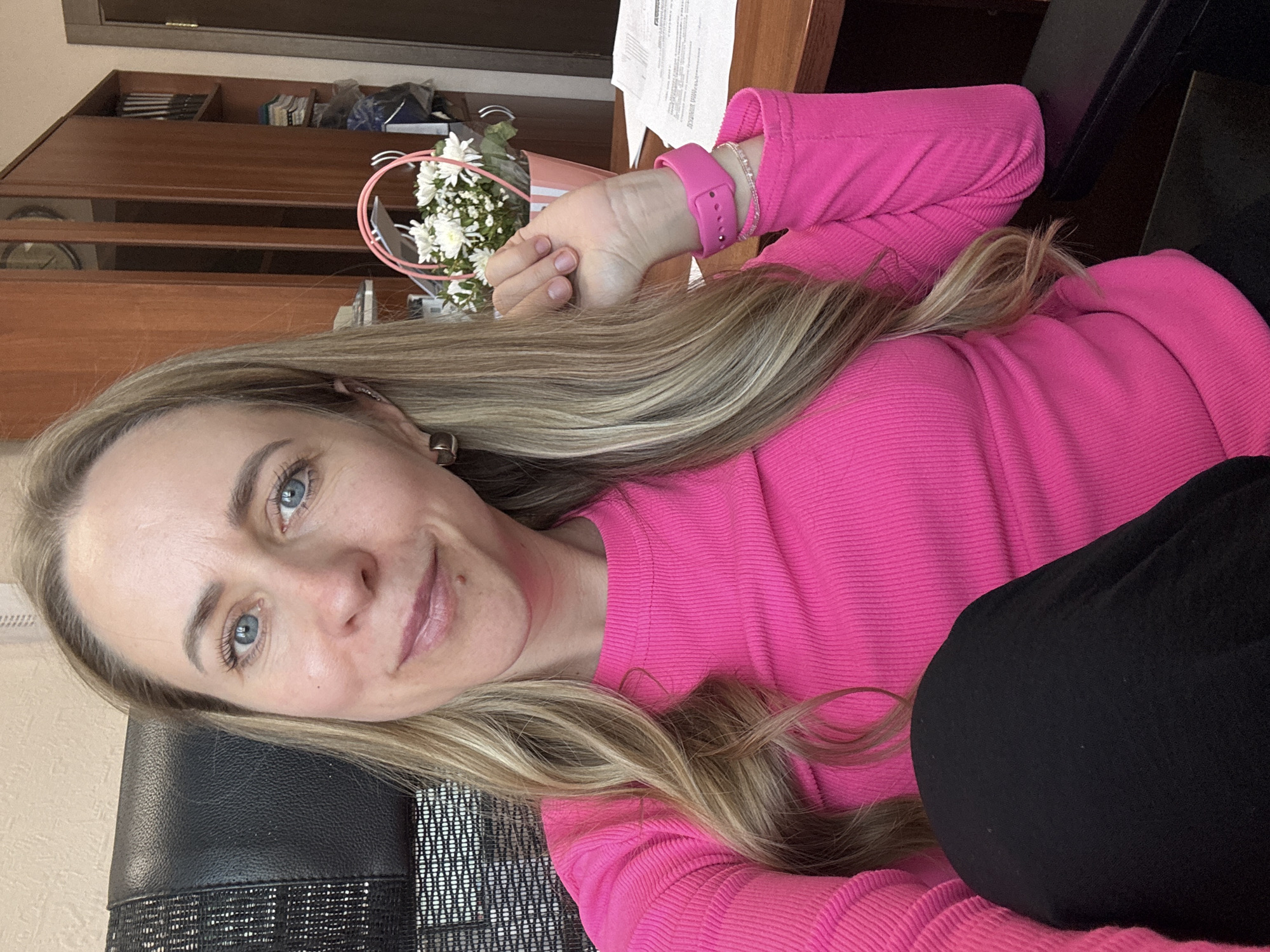 A woman with fair skin, blonde hair, and blue eyes is posing for a selfie. She is wearing a bright pink long-sleeved shirt and a pink smartwatch. She is seated on a black office chair. In the background, there is a wooden cabinet, a desk with papers, and a vase of white flowers. The location is likely an office setting in Chaikovsky, Russia.