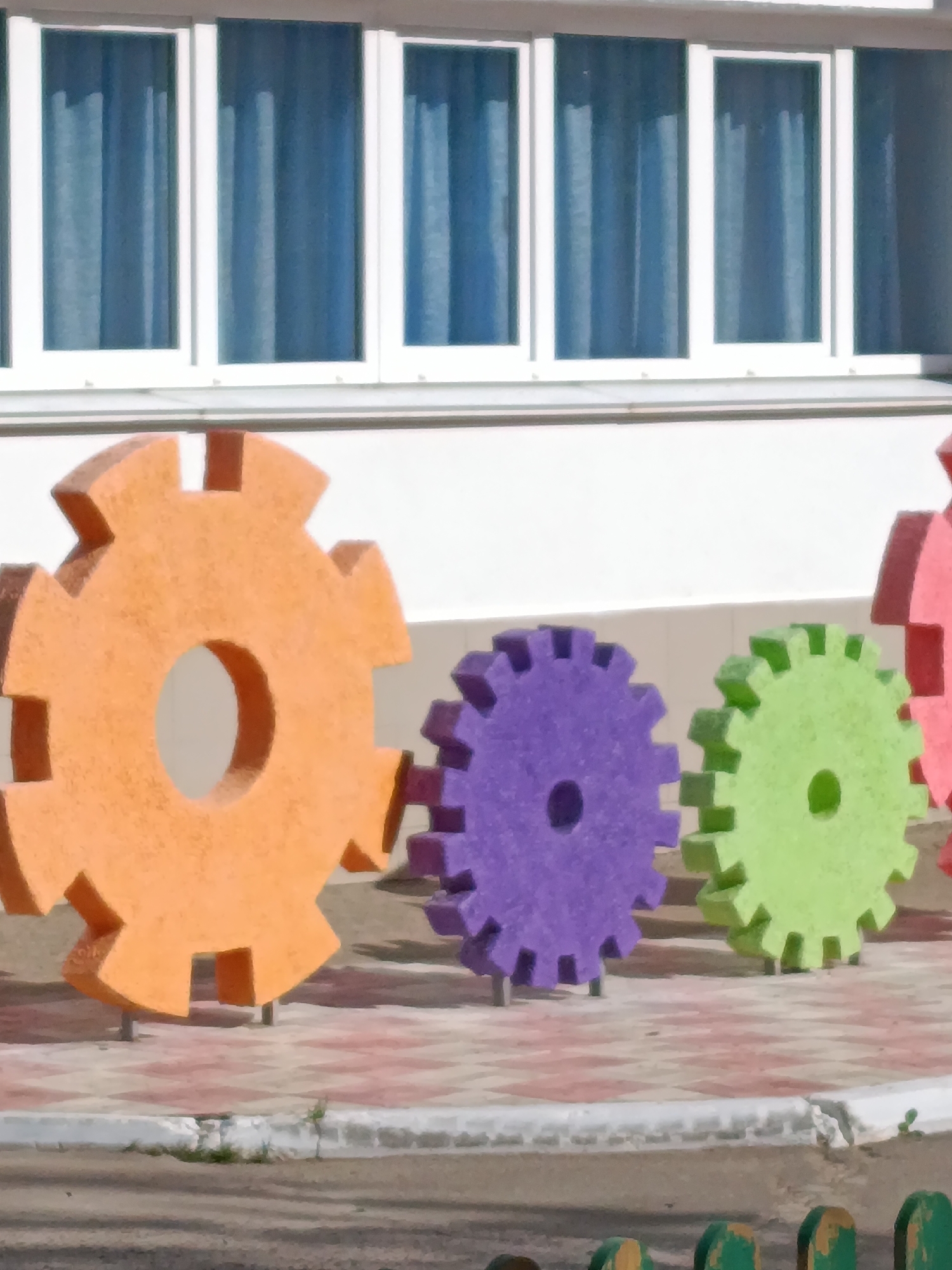 The image shows a row of oversized, colorful gear-shaped sculptures in Chaikovsky, Russia. The gears are orange, purple, green, and red. They are located on a paved area with a light-colored concrete border. Behind the gears is a white building with windows that have blue curtains. The lower part of a green and brown fence is visible in the foreground.