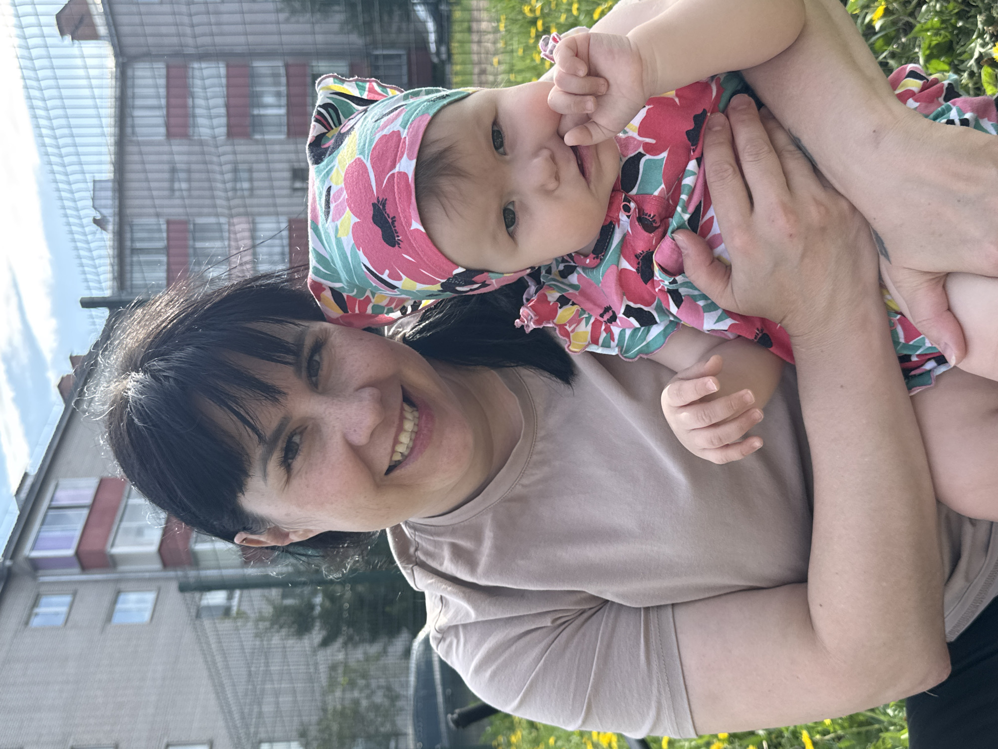 A woman with dark hair and bangs holds a baby in a colorful floral dress and head covering. Both are smiling and looking at the camera. The woman wears a tan t-shirt. They are outdoors, likely in a residential area, with a building visible in the background that has a light-colored facade and red window frames. Yellow flowers grow in the foreground. The image appears to be taken on a sunny day. The location is Zav'yalovo, Russia.
