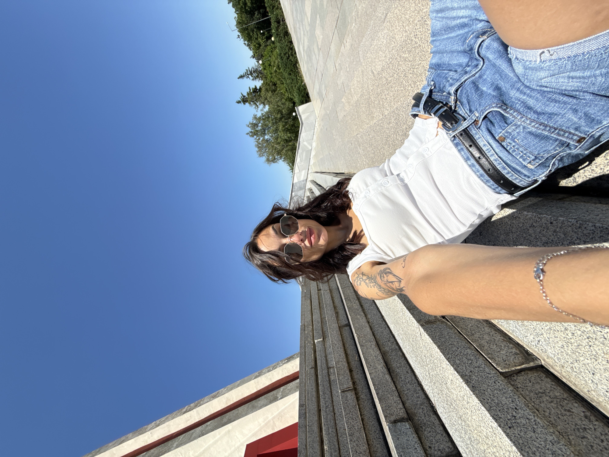 A young woman with dark hair and sunglasses is taking a selfie while sitting on wide, stone steps outdoors. She is wearing a white button-up top and denim shorts. A tattoo is visible on her left arm, and a silver bracelet is on her wrist. Behind her, a large, modern structure with red and white accents is visible. The sky is clear and blue, and the overall environment suggests a bright, sunny day. The location is the Monument of the Liberators – Pleven Panorama, located in the city of Pleven, Bulgaria.