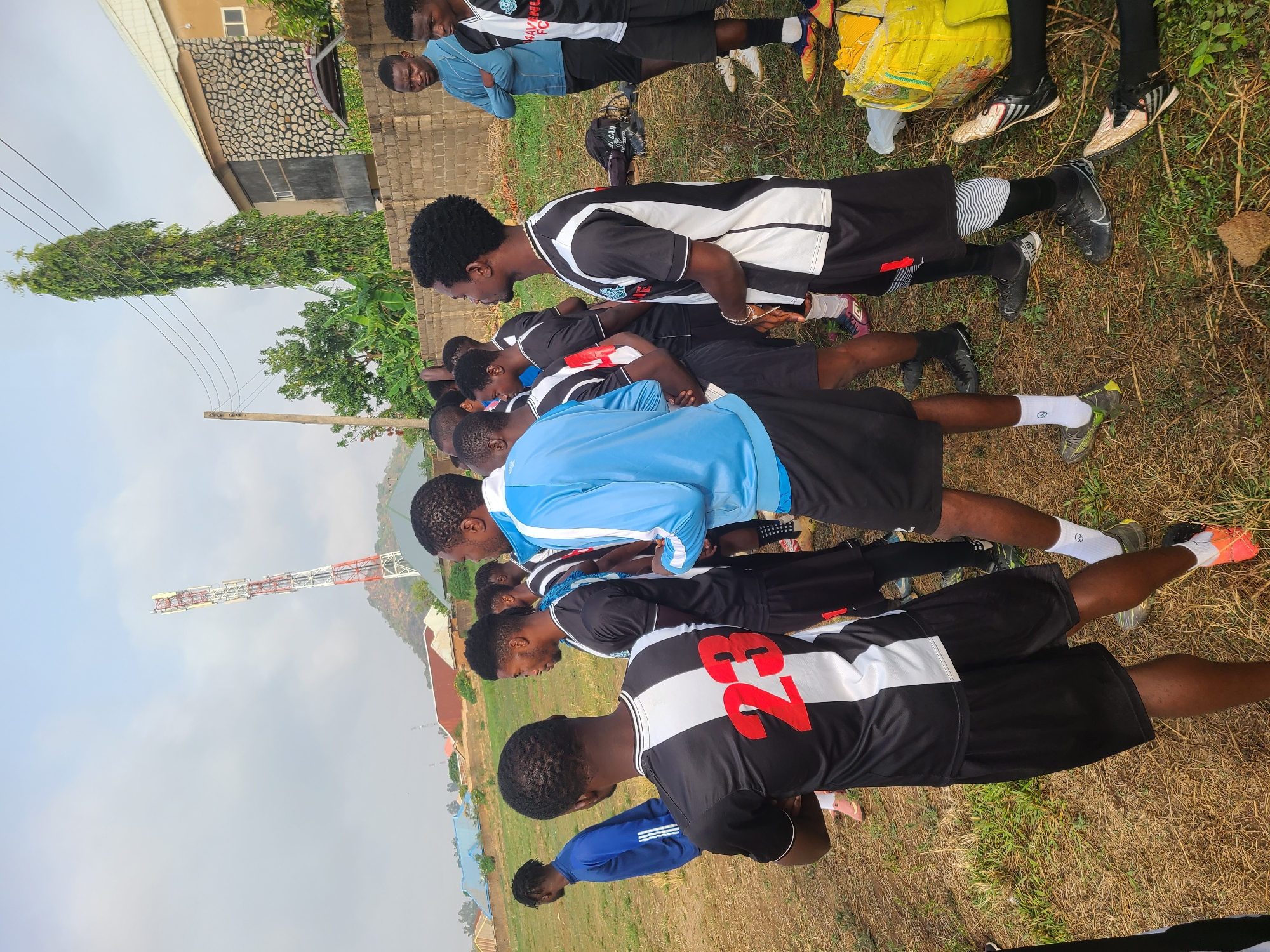 The image shows a group of approximately ten young men standing in a row on a grassy field in Bukuru, Nigeria. They appear to be soccer players, wearing black and white jerseys with numbers. Some have on soccer cleats. The weather is overcast. In the background, there is a building, a communication tower, trees, and a hill.