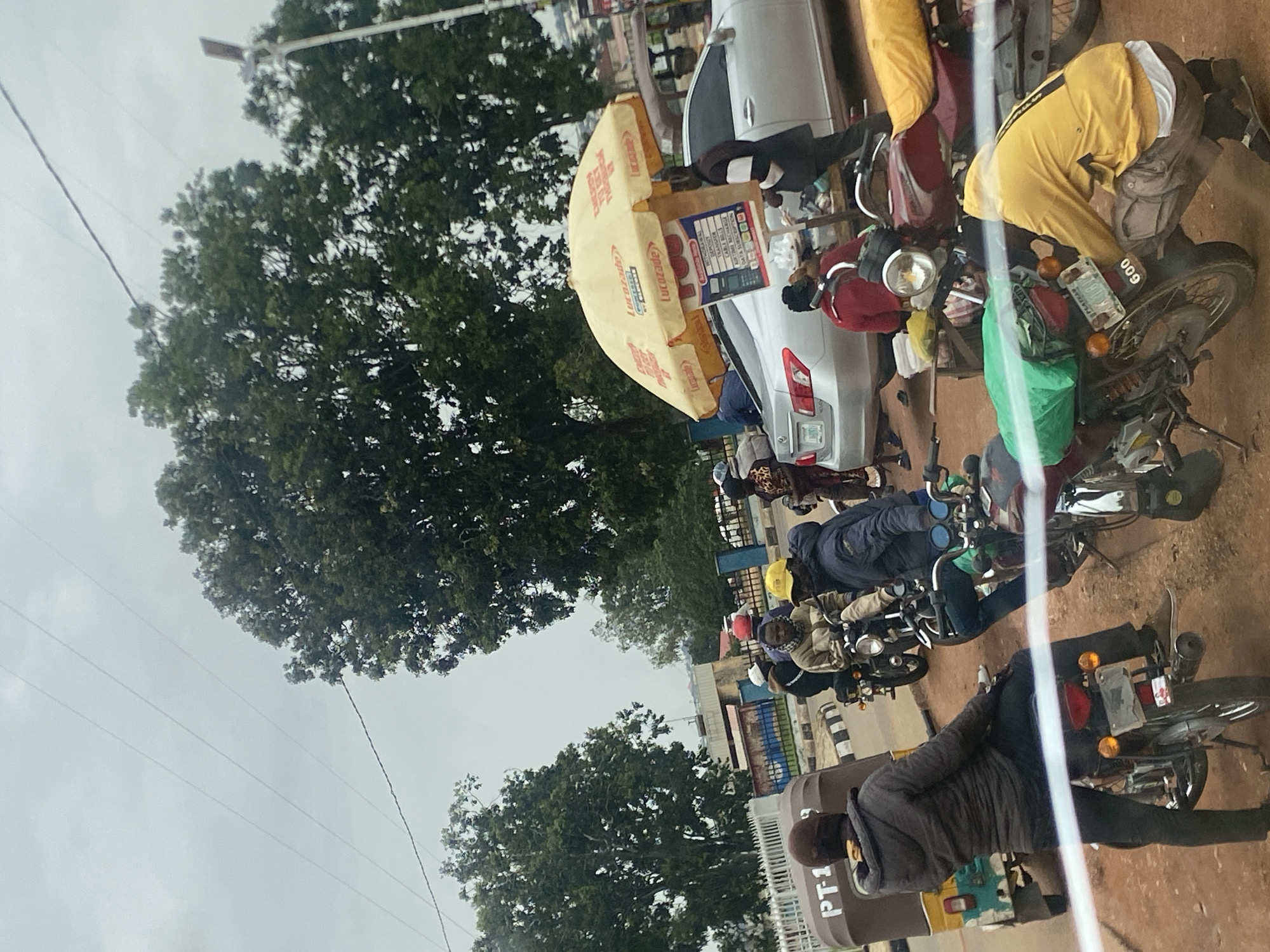 This image depicts a busy street scene in Gura Top, Nigeria, likely during the daytime given the ambient light. Several motorcycles are parked along the side of the road, with a few people attending to them or interacting with a vendor. One prominent object is a yellow umbrella with "Lucosade" branding, shading a small stall where items are displayed, including what appears to be a calculator and some papers.
People are seen in various casual outfits, some wearing jackets and beanies, suggesting cooler weather or a personal preference. One individual in the foreground wears a grey jacket and black pants, while another person in the background is dressed in bright yellow trousers. The overall atmosphere seems active, with individuals engaged in commerce or transit. The presence of motorcycles indicates transportation is a key element of the scene. The ground is a reddish-brown, typical of dirt or unpaved roads. Trees with lush green foliage line the background, and power lines are visible overhead, stretching across the frame. The sky appears overcast, hinting at a cloudy day. Some text is visible on the yellow umbrella, as well as on a small sign on the stall and on the back of one of the motorcycles, which appears to say "009". There is also partial text on a vehicle in the lower left that reads "PT-1".