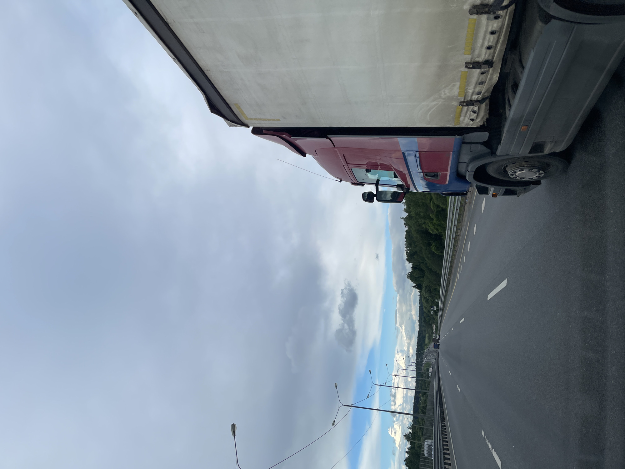 A truck is positioned on a highway near Бавыкино, Russia. The truck is red and blue. The sky is cloudy. The highway has multiple lanes and white dashed lines. Streetlights line the side of the road. Trees are visible in the background.