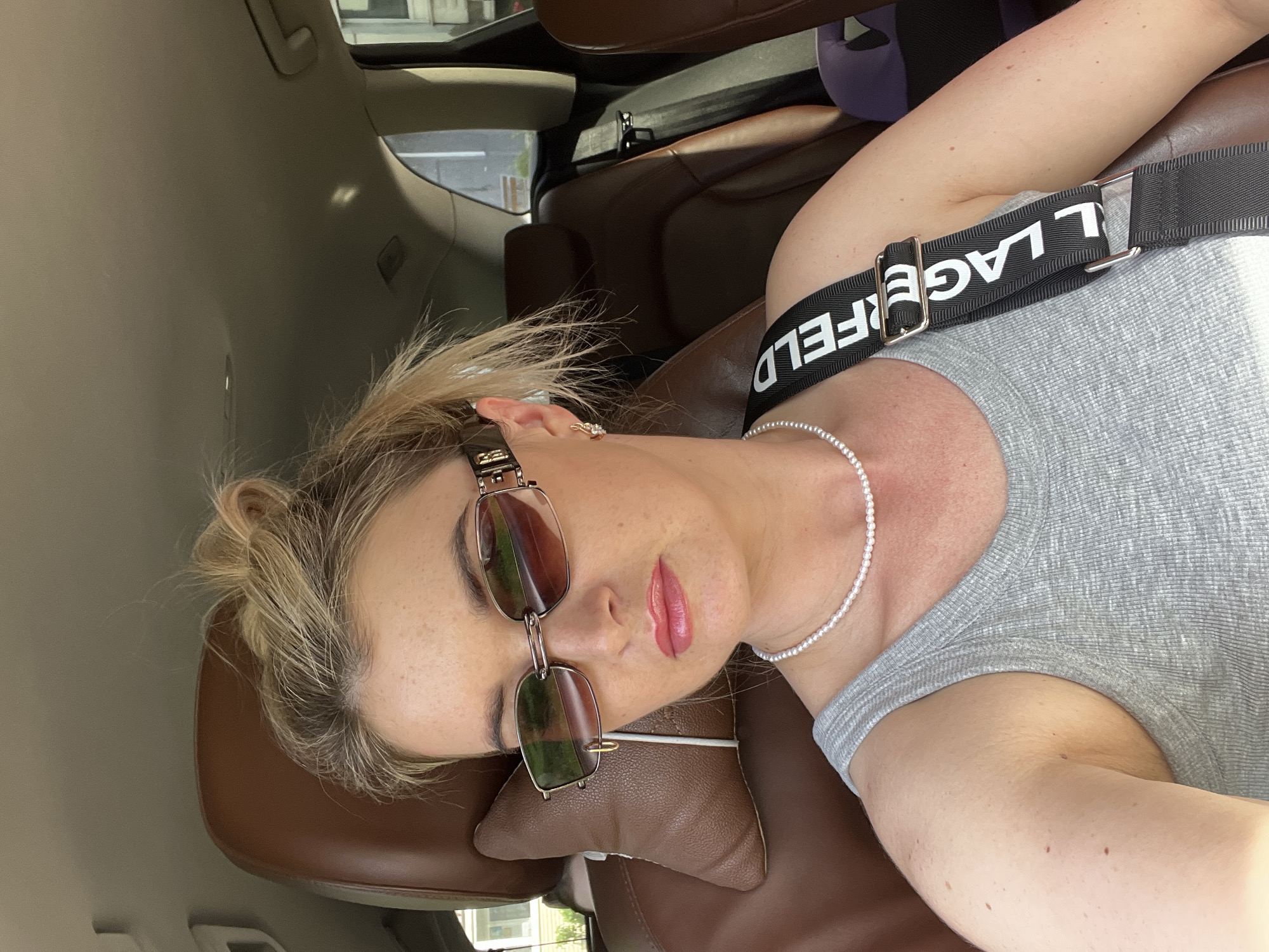 A blonde-haired woman with freckles is positioned in the passenger seat of a car, possibly a brown leather-seated SUV. She wears sunglasses, a pearl necklace, and a gray tank top with a Karl Lagerfeld-branded shoulder strap. The location, Chaikovsky, Russia, is not definitively confirmed by visual cues within the image. The scene depicts a moment of rest or relaxation within a vehicle, but further details are limited.