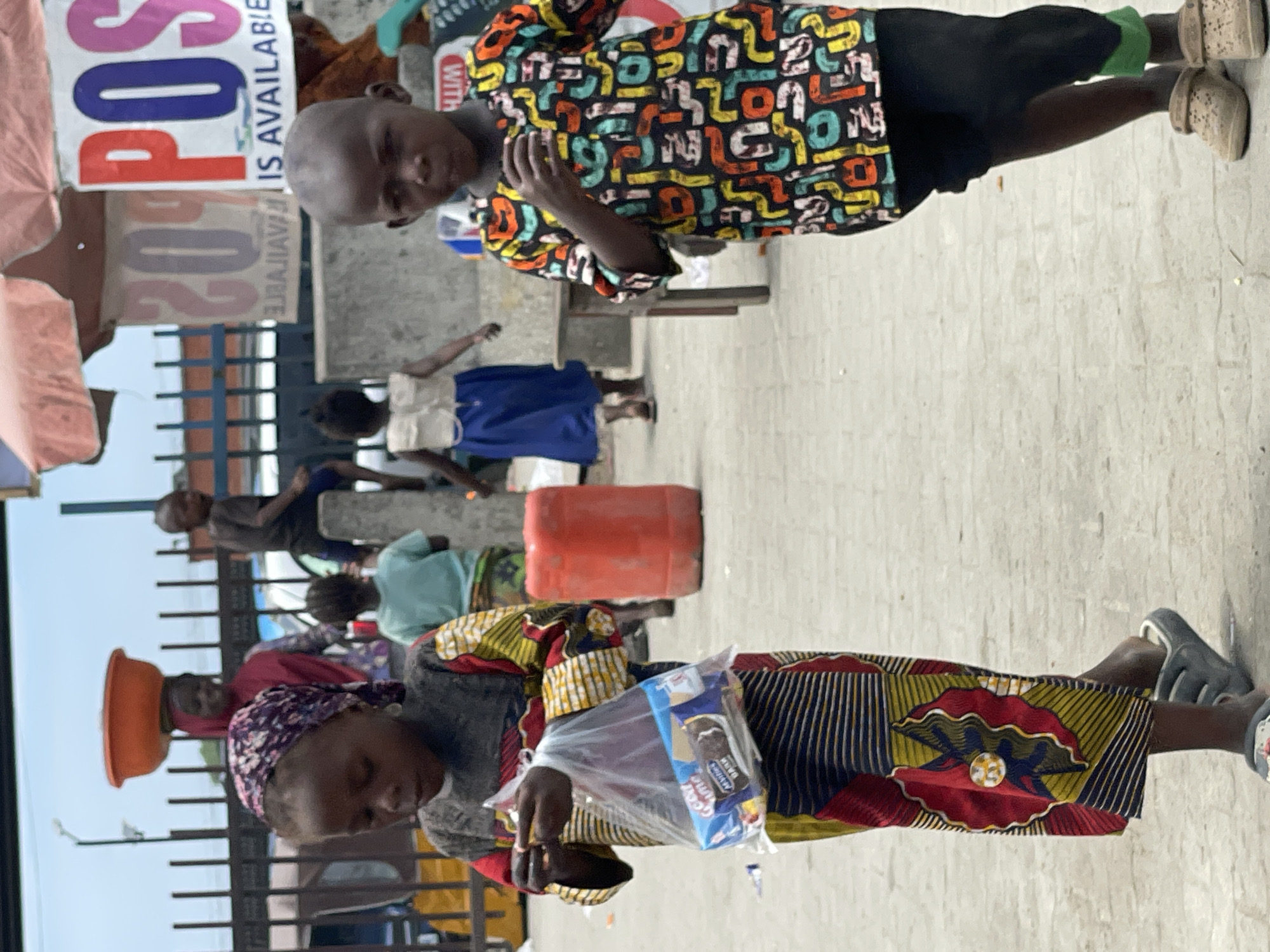 The image depicts a street scene in Lekki, Nigeria. A woman in a colorful patterned dress and headscarf holds a plastic bag containing packaged snacks. Nearby, a child in a patterned shirt and black pants stands facing the viewer. Other people and children are visible in the background. A red plastic container sits on the ground, and a sign reading "POS IS AVAILABLE!" is partially visible. The environment appears to be a paved area along a street with buildings.