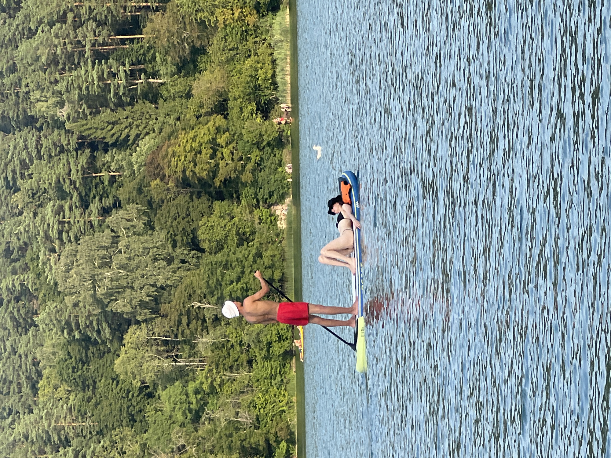 In the city of Vilnius, Lithuania, on a body of water surrounded by a dense forest of green trees, a young man stands on a paddleboard, holding a paddle and wearing red shorts and a white hat. He is facing away from the viewer. Beside him, a young woman lies on an inflatable raft, wearing a black bikini and a black hat. The water is rippled and reflects the blue sky. In the background, several other people are visible on the water and on the shore. A bird is flying over the water. The scene appears to be a recreational setting.