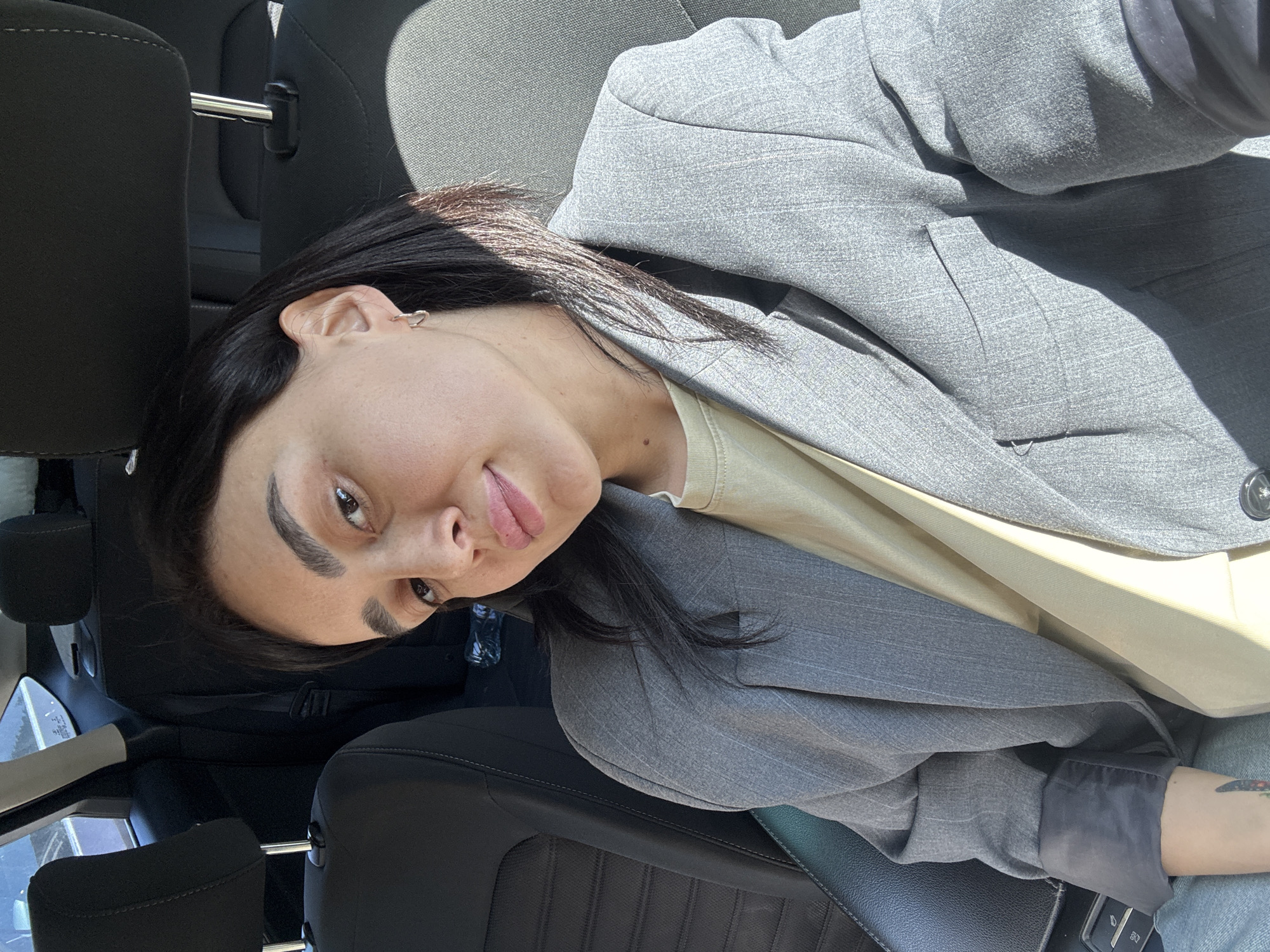 The image shows a person sitting in the driver's seat of a car. The person has dark hair, fair skin, and is wearing a grey blazer over a light-colored shirt. The car's interior is black with grey accents. The background suggests a sunny day, but no specific landmarks are visible to confirm a location in Tbilisi, Georgia. The person is looking at the camera. A headrest is visible behind them. The angle of the shot suggests it was taken from above or slightly to the side of the person.
