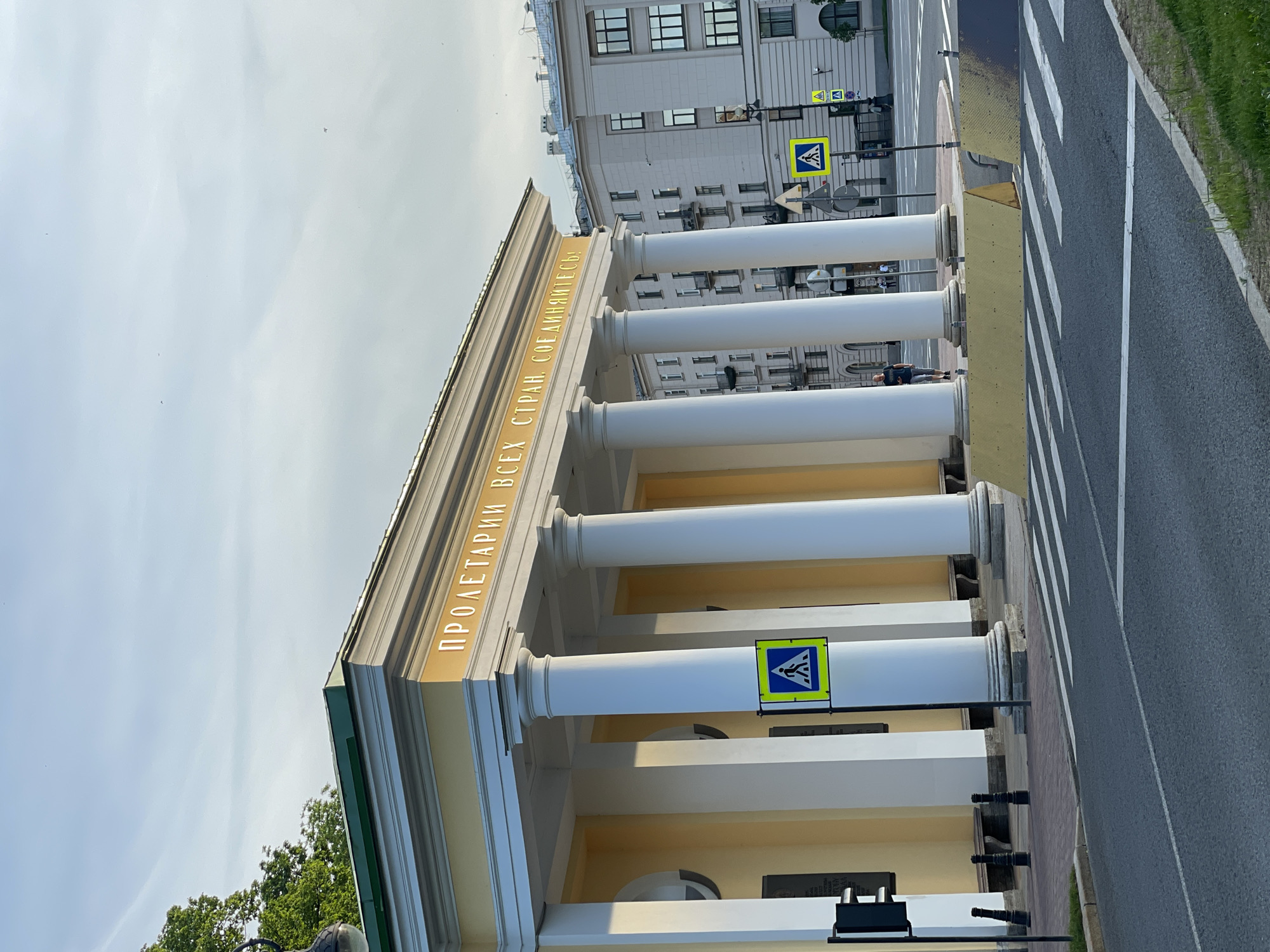 The image depicts the Narva Triumphal Arch in Saint Petersburg, Russia, under a partly cloudy sky. The monumental yellow-painted structure, featuring a green roof and a series of prominent white Doric columns, is oriented towards the viewer's left. Along its frieze, the gold Cyrillic inscription "ПРОЛЕТАРИИ ВСЕХ СТРАН, СОЕДИНЯЙТЕСЬ!" (Proletarians of all countries, unite!) is visible.
In the foreground, a two-lane asphalt road with a marked white-striped pedestrian crossing extends horizontally. A narrow strip of green grass borders the road on the immediate right. On the far side of the road, two yellow, wedge-shaped barriers are placed on the sidewalk. A black traffic light pole with a signal head stands on the left side of the pedestrian crossing. Near the building, a prominent blue and yellow pedestrian crossing sign is mounted on a pole, with other signs partially visible behind it. Black bollards line the sidewalk in front of the arch.
One adult male, wearing a dark t-shirt and shorts, walks on the sidewalk in front of the arch's columns, moving from right to left. Additional blurred figures are faintly visible in the background on the far right. Behind the arch and to its right, multiple-story urban buildings with numerous windows and balconies are visible under the same sky. The lighting suggests natural daylight, casting shadows that illuminate the arch's facade.