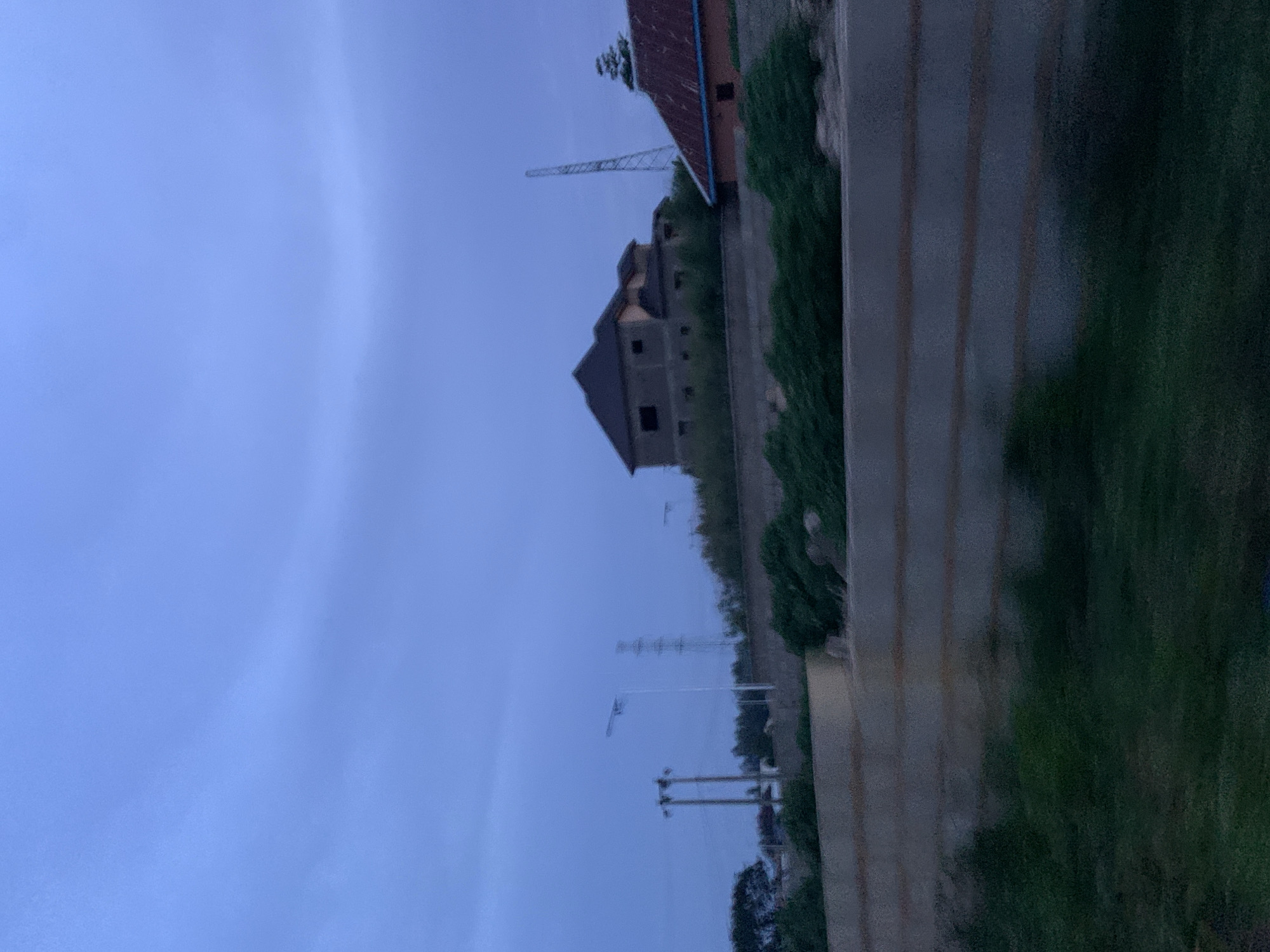 The image shows a twilight scene in Gura Top, Nigeria. A large, unfinished concrete house with dark windows stands on a hill, partially obscured by lush green foliage. To its right, a smaller building with a red corrugated roof is visible. In the background, a tall, slender transmission tower pierces the hazy sky, and two power lines are also present. The foreground features a textured concrete embankment and dark green grass. The sky is a muted blue, indicating the transition from day to night.