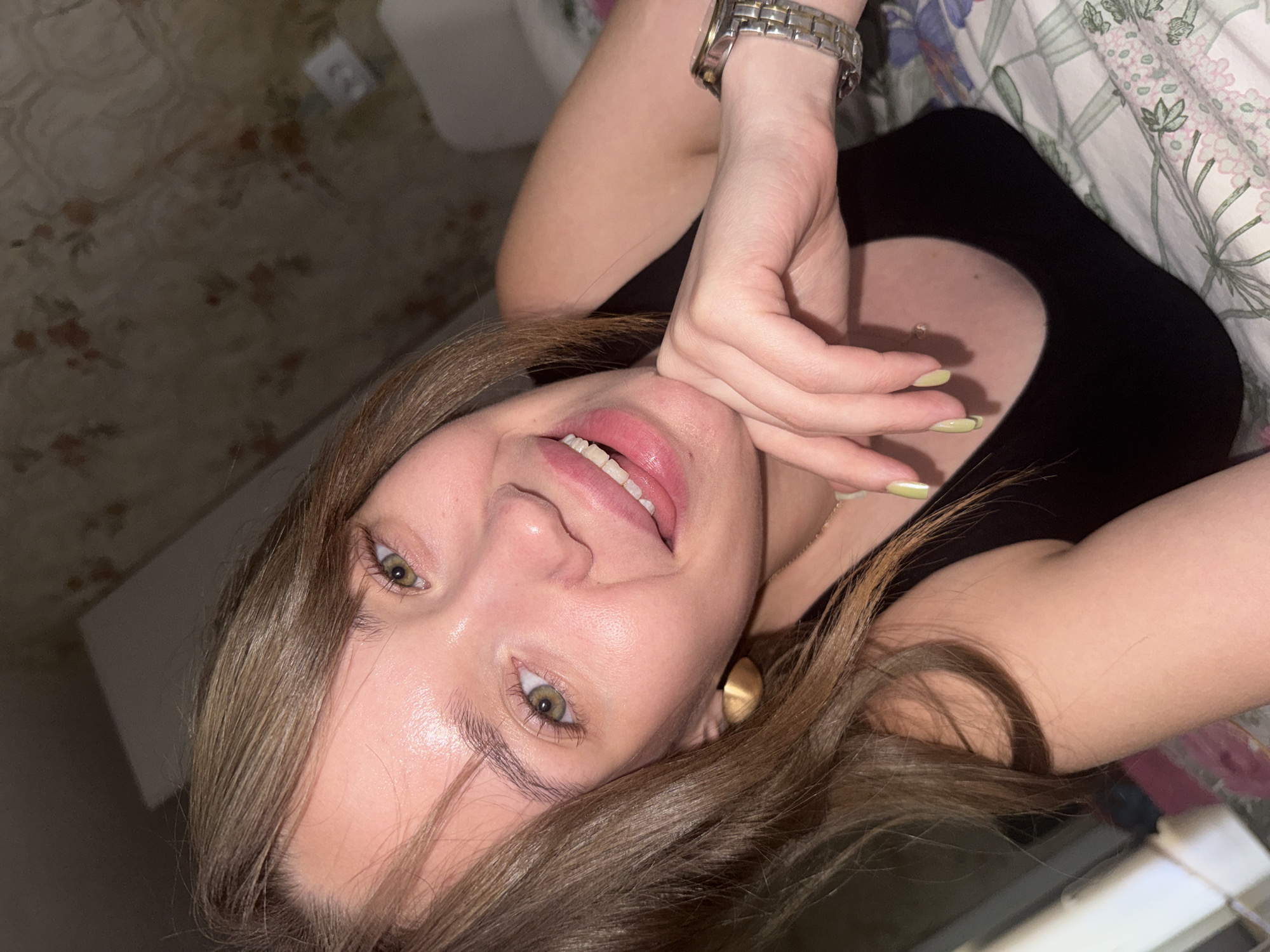 A young woman with brown hair and green eyes is captured in a close-up, tilted selfie, likely taken in her bedroom in Chaikovsky, Russia. She is wearing a black tank top and has her tongue sticking out playfully. Her left hand is resting on her chin, and a silver watch is visible on her wrist. Her fingernails are painted a light green color. The background shows a floral patterned wallpaper and a bed with a floral comforter. The lighting is dim, suggesting the photo was taken indoors, possibly at night or in a low-light environment.