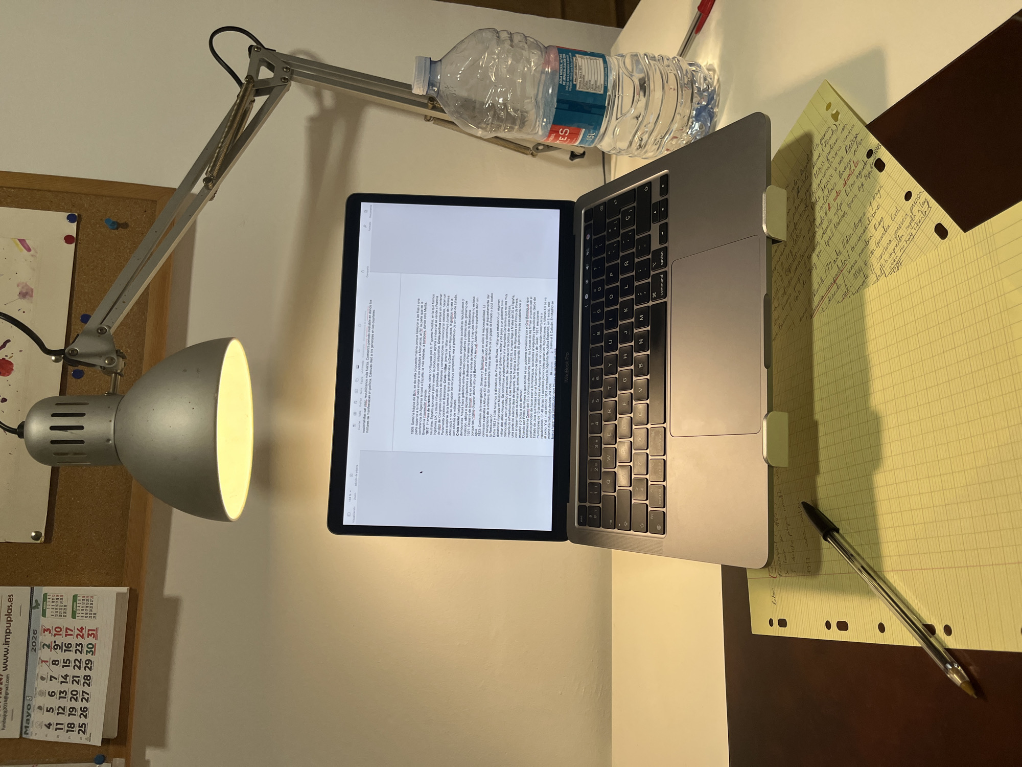 Here's a description of the image:
The image shows a desk illuminated by an adjustable lamp. A silver laptop sits open on the desk, displaying a document in Spanish. To the right of the laptop are yellow lined notes with handwritten text, and a pen. A bottle of water sits near the top of the laptop. To the left of the laptop, a corkboard is attached to the wall, along with a calendar. The calendar month is May 2026. The location, Torrent, Spain is not detectable.